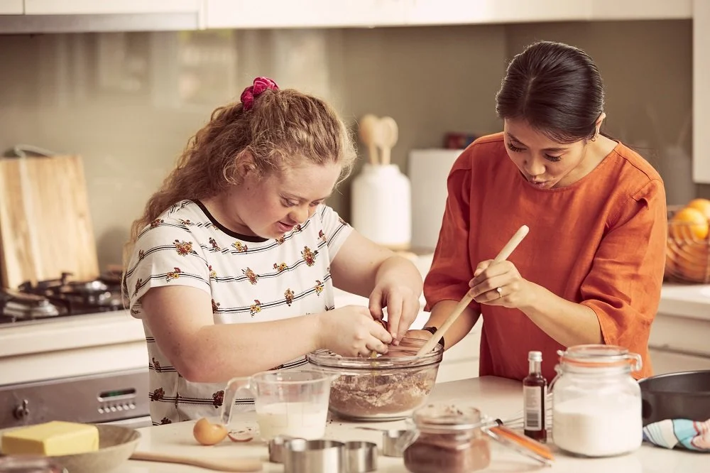 A woman and a girl baking together in a kitchen. The girl is mixing ingredients in a glass bowl while the woman assists.
