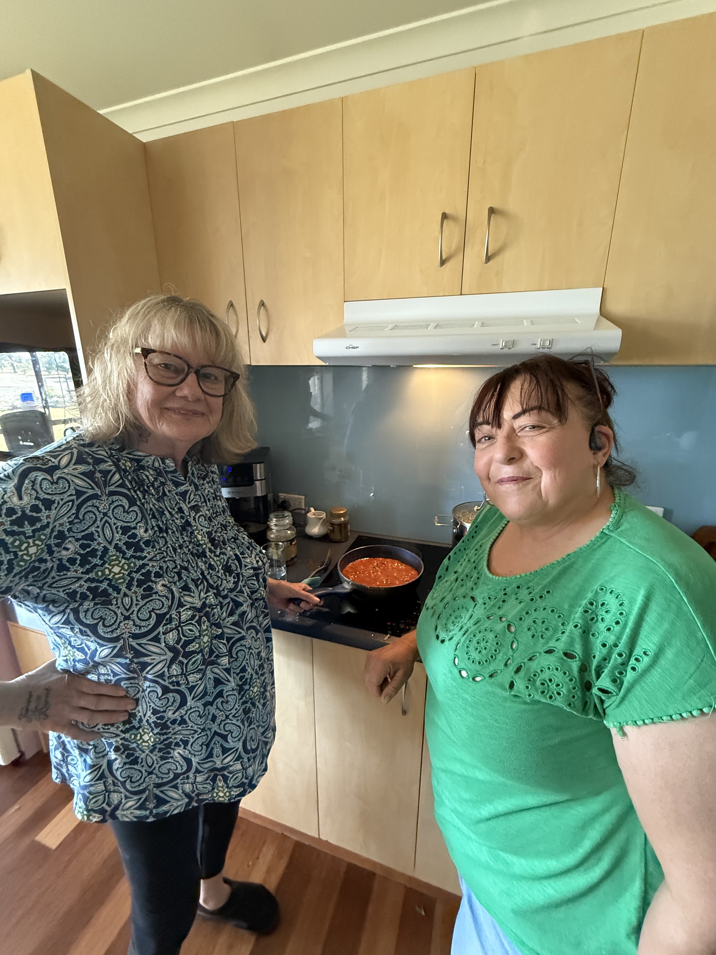 Two women cooking together in a kitchen, one holding a frying pan with tomato sauce, smiling at the camera.