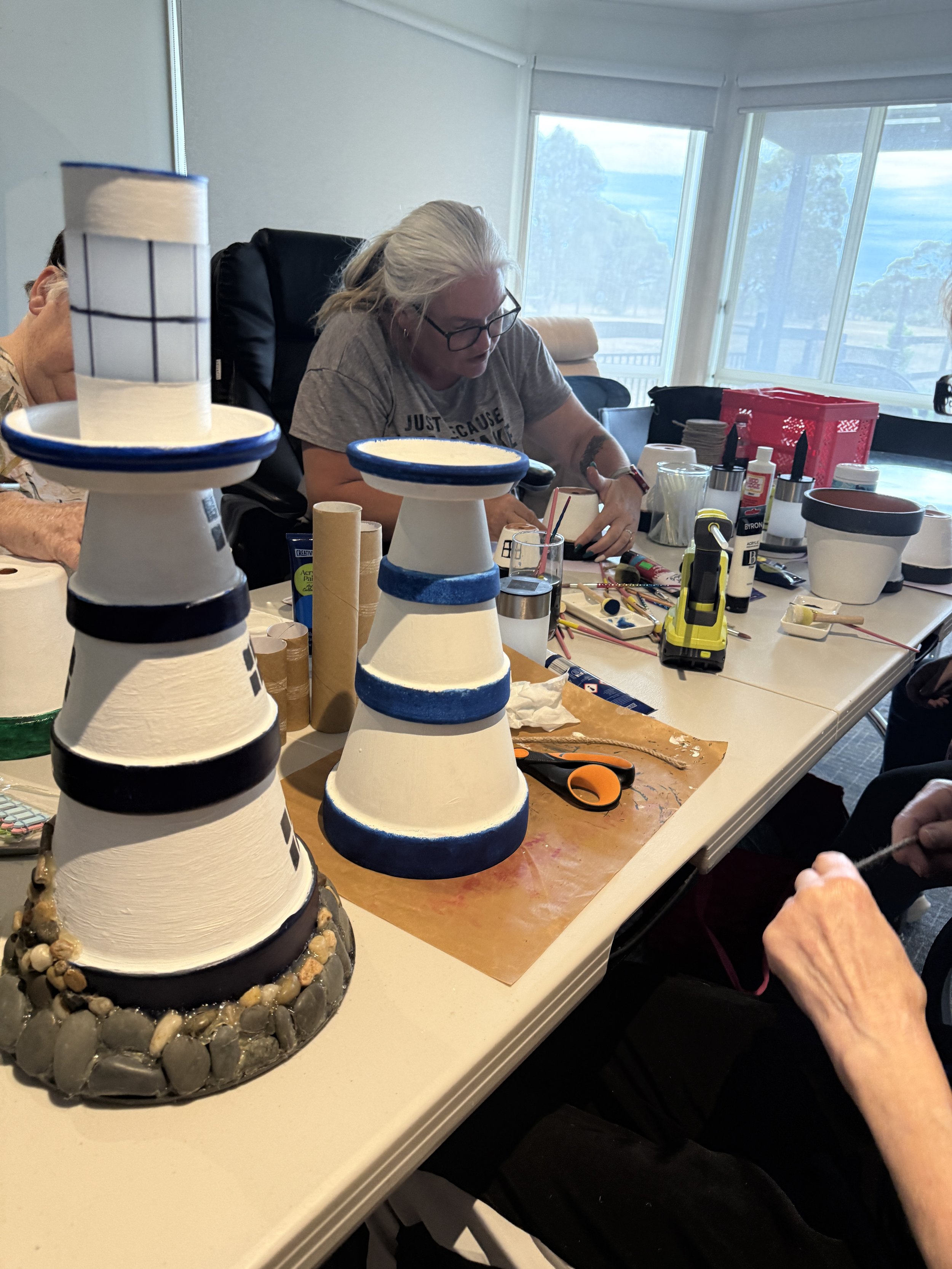 People sitting around a table working on craft projects, with painted stacked ceramics and various supplies such as scissors, glue, and paint markers.