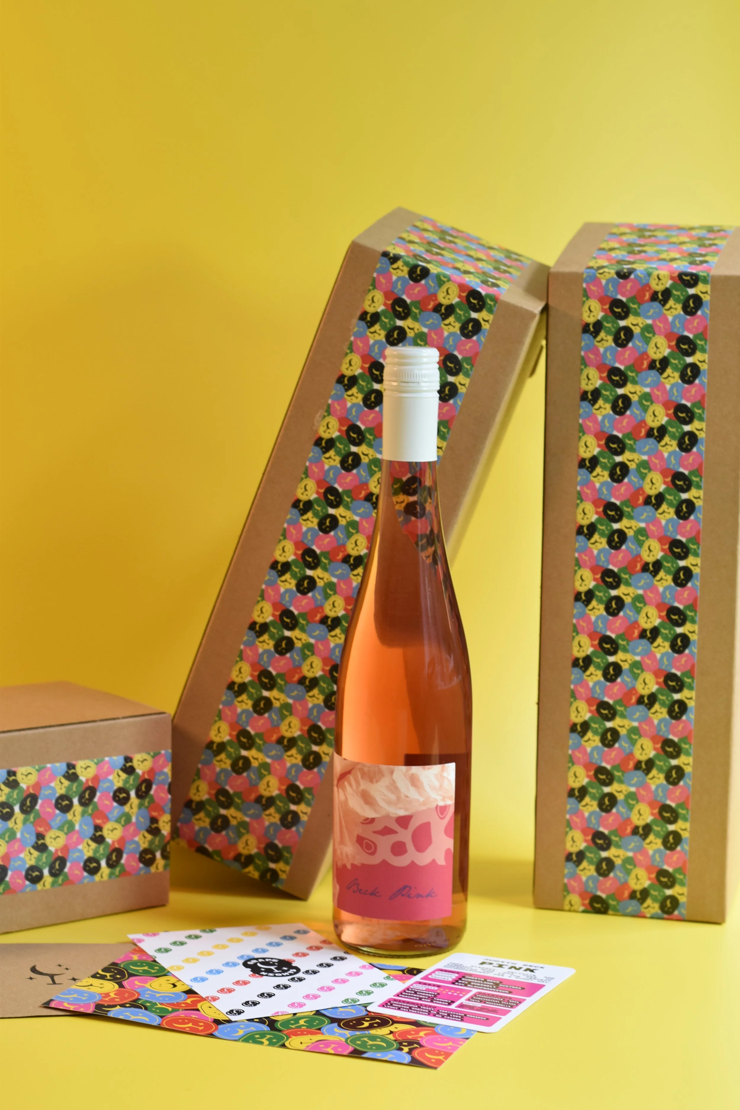 A pink rosé wine bottle with a white cap and label, featuring abstract pink shapes, positioned among gift boxes and sheets of colorful smiley face stickers, all set against a yellow background.