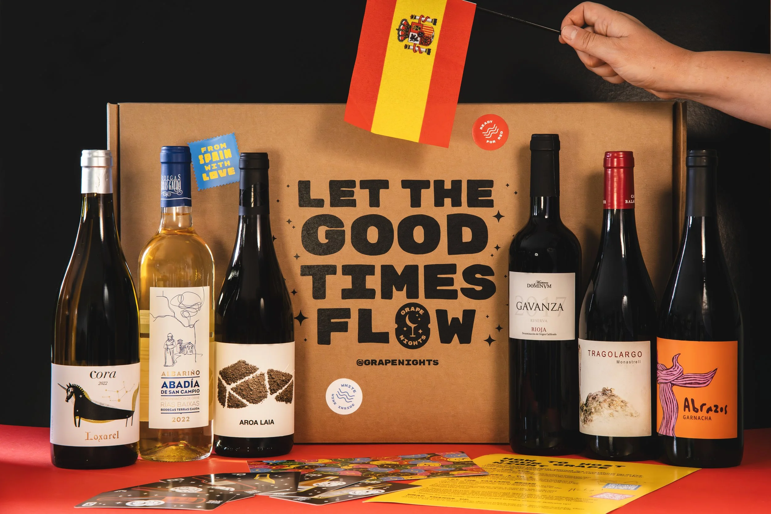 A box with the message 'Let the Good Times Flow' is surrounded by six bottles of wine, each with unique labels, against a black background. A hand is holding a small Spanish flag above the box, and promotional cards and a yellow flyer are on the red