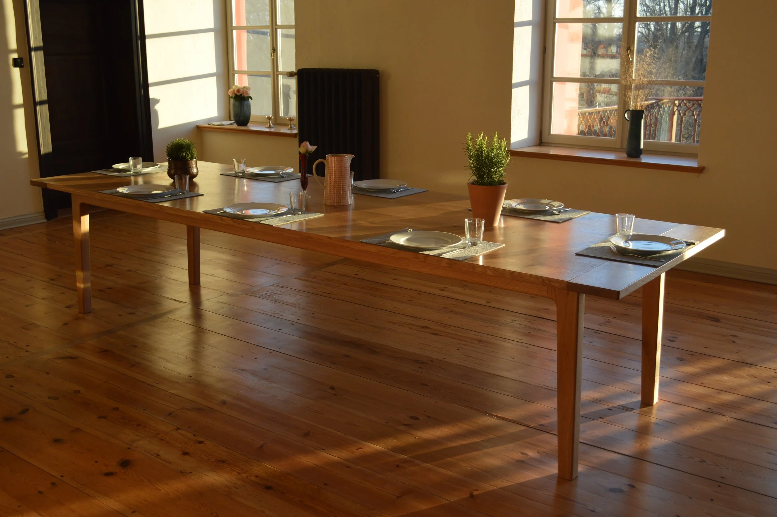 Ein heller Speisesaal mit einem langen Holzlagertisch, gedeckt mit Tellern, Gläsern, einer Vase mit Blumen und Topfpflanzen, Sonnenlicht durch große Fenster
