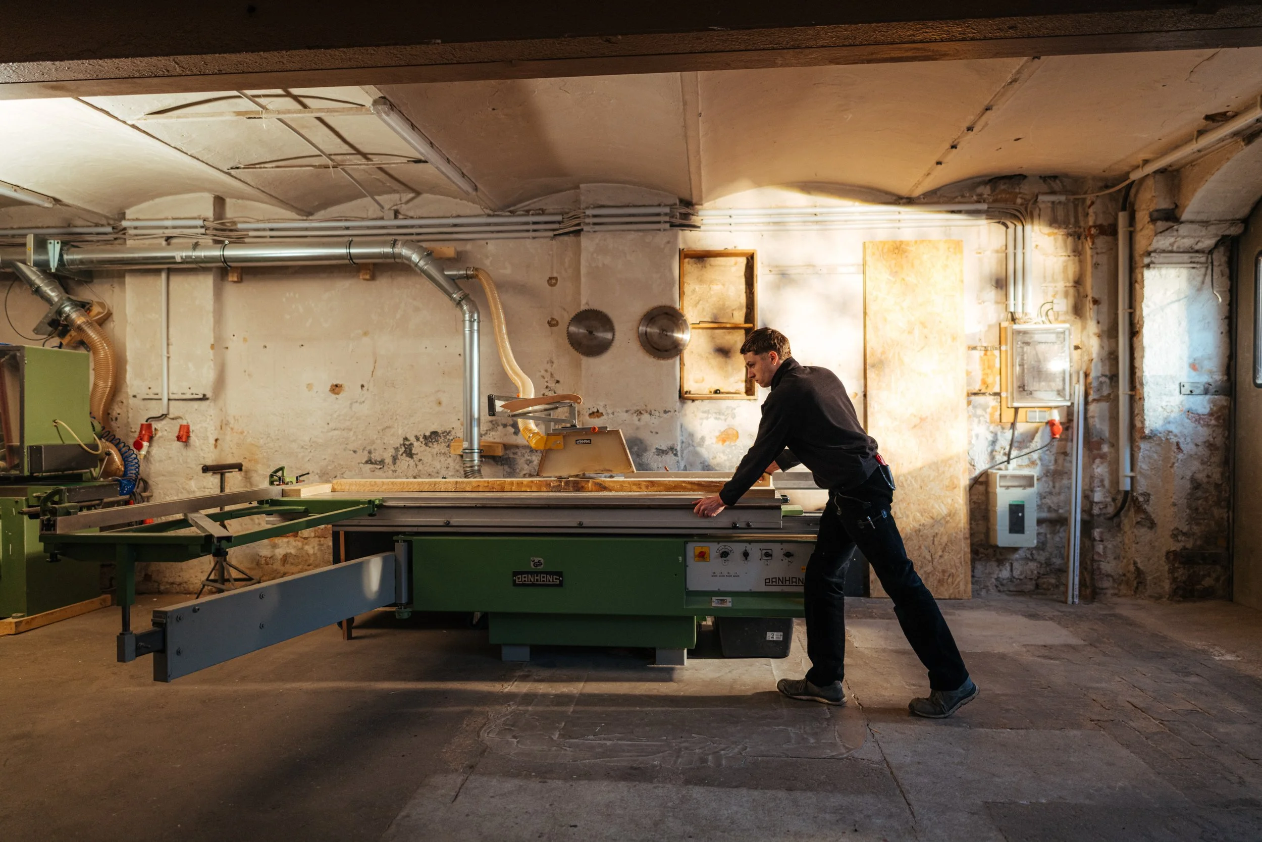Mann arbeitet mit einer Tischsäge in einer Werkstatt mit roher Wand und Installationen im Hintergrund.