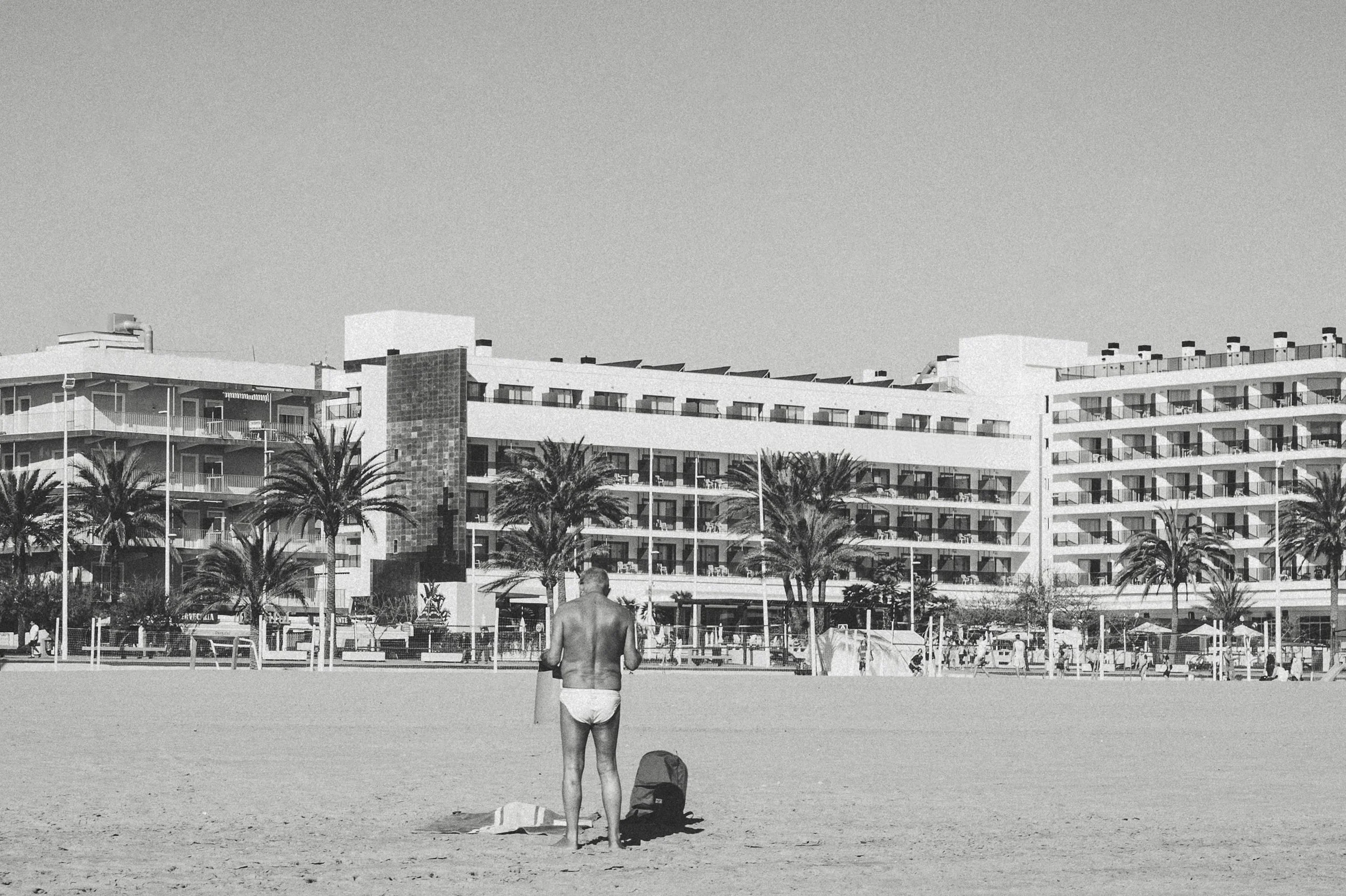 En man i badkläder står på stranden med rygg mot kameran, framför ett hotellkomplex med palmträd.