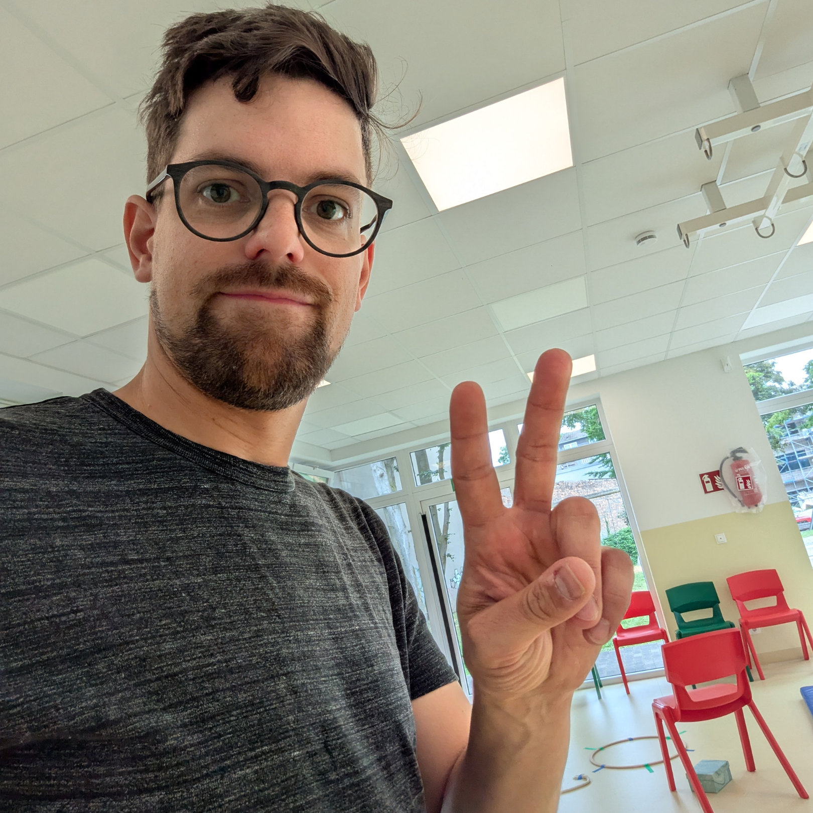 Thomas trägt eine schwarze Brille und macht ein Peace-Zeichen mit der Hand, im Hintergrund sind farbige Stühle und große Fenster in einem Raum. Lass uns gemeinsam was bewegen!