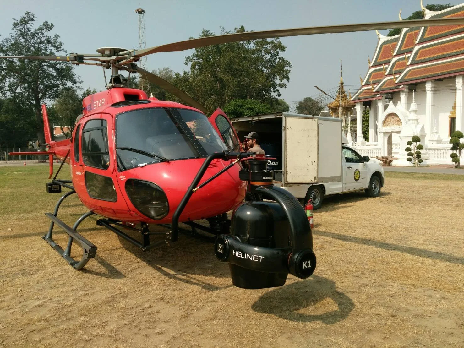 A red helicopter parked on a grassy area with a white vehicle and traditional Thai-style building in the background.