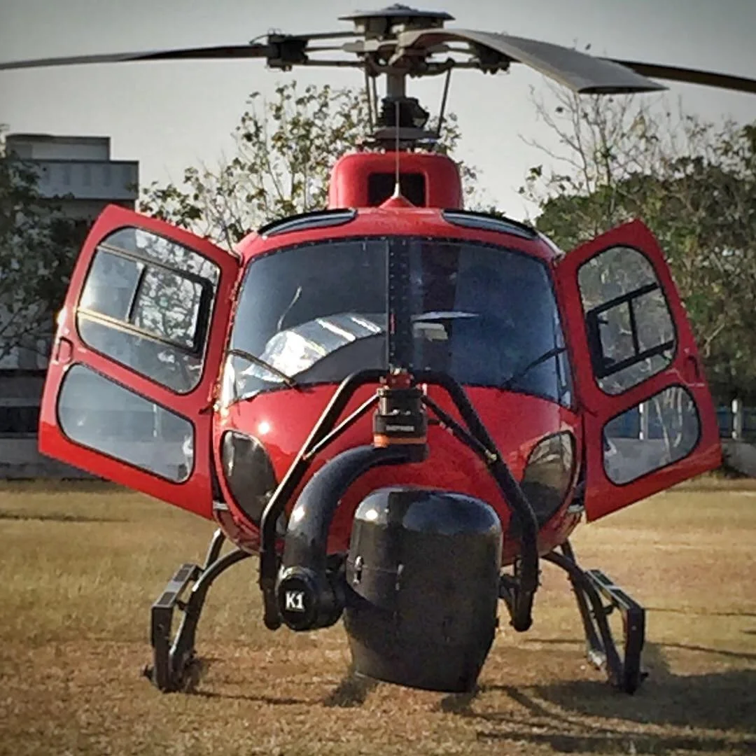 Front view of a red helicopter with open cockpit doors, parked on grass with trees and a building in the background.