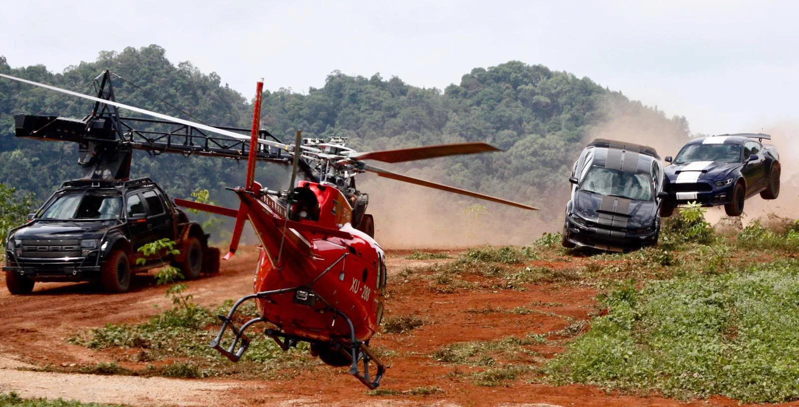 A black Ford vehicle is in a midair collision with a blue and black sports car, while a red helicopter hovers nearby on a dirt path surrounded by greenery and hills.