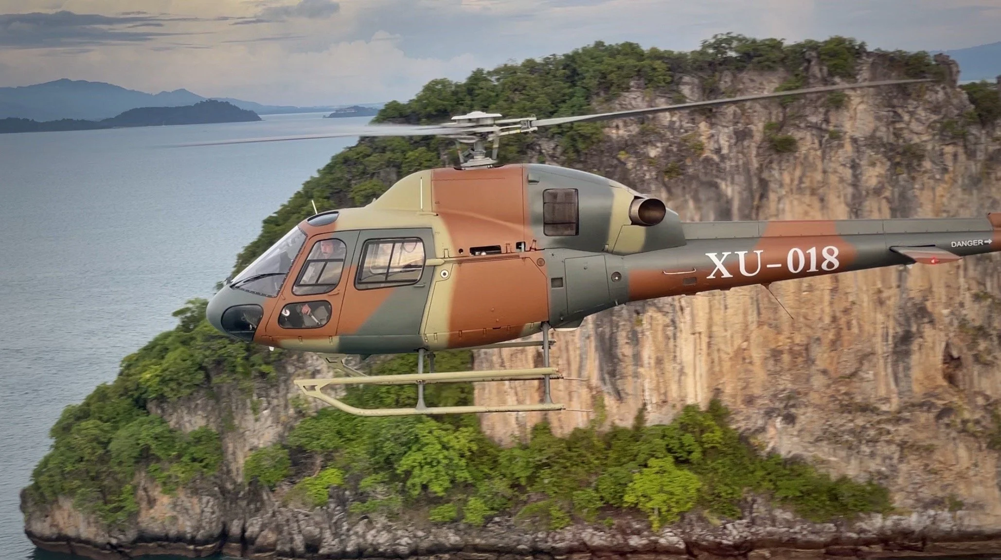 A military helicopter flying over a body of water with a rocky, green-covered island in the background.