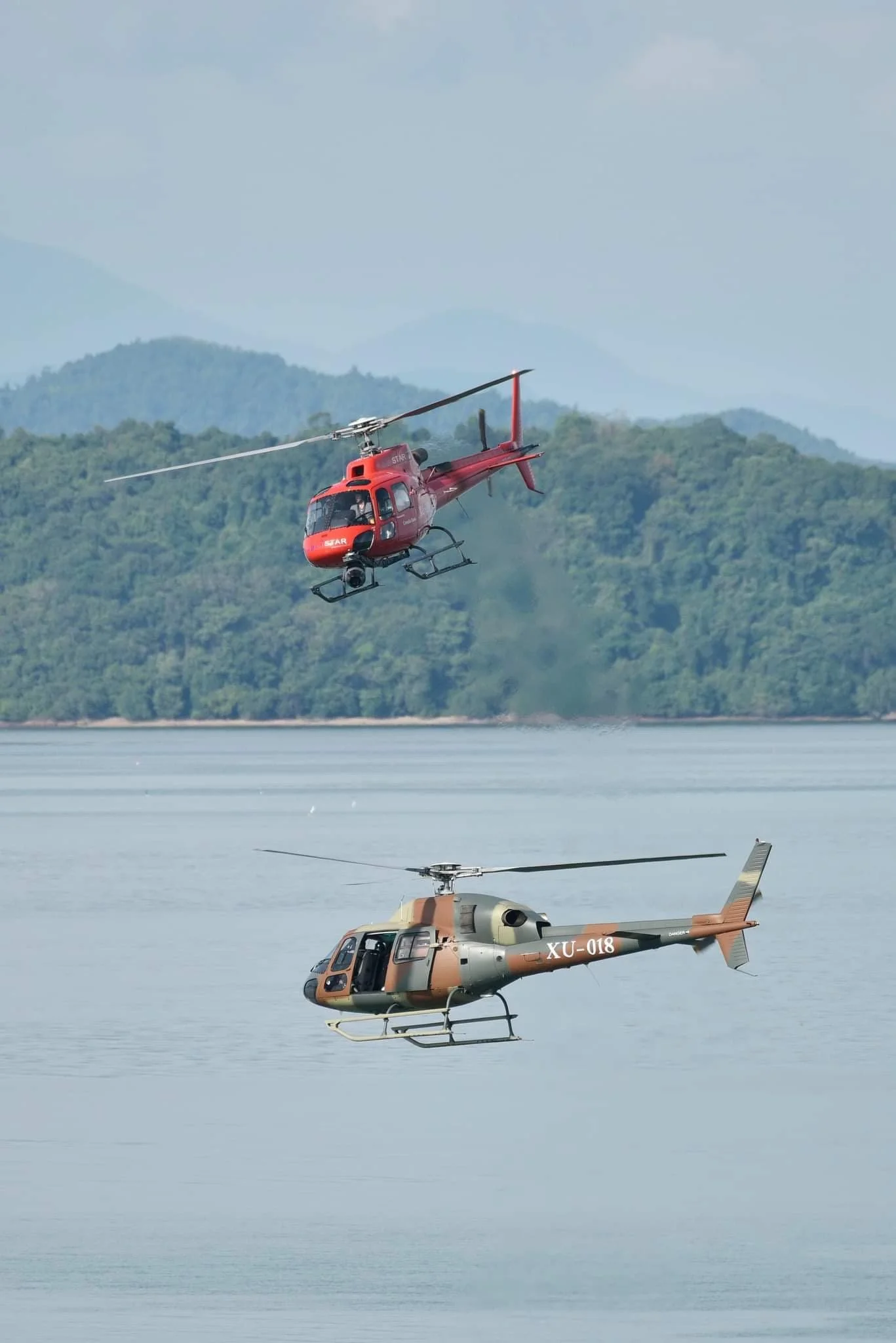 A red helicopter flying above a body of water with another helicopter floating just above the water surface, with a forested mountain range in the background.