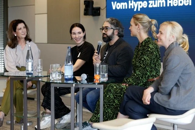 Fünf Menschen sitzen an einem Tisch bei einer Podiumsdiskussion oder Fragerunde, mit Wasserflaschen und Gläsern auf dem Tisch, im Hintergrund ein Bildschirm mit dem Text 'Vom Hype zum Hebel'.