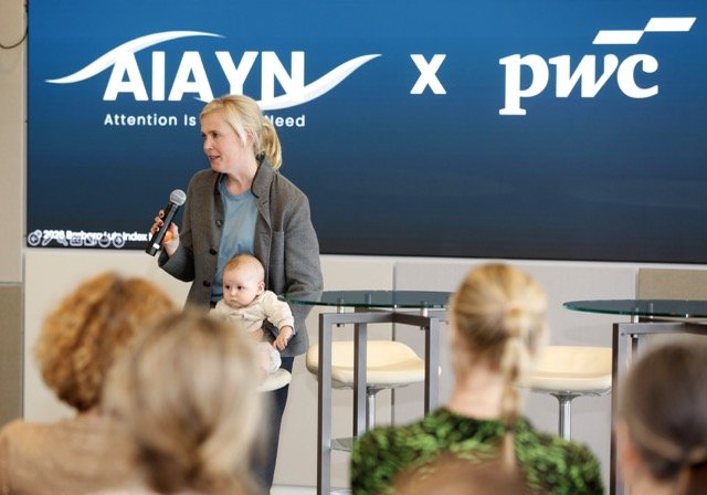 Eine Frau hält ein Baby im Arm und spricht bei einer Veranstaltung. Im Hintergrund sind Logos von AIYN und PwC auf einem Bildschirm zu sehen, während im Vordergrund mehrere Menschen sitzen.