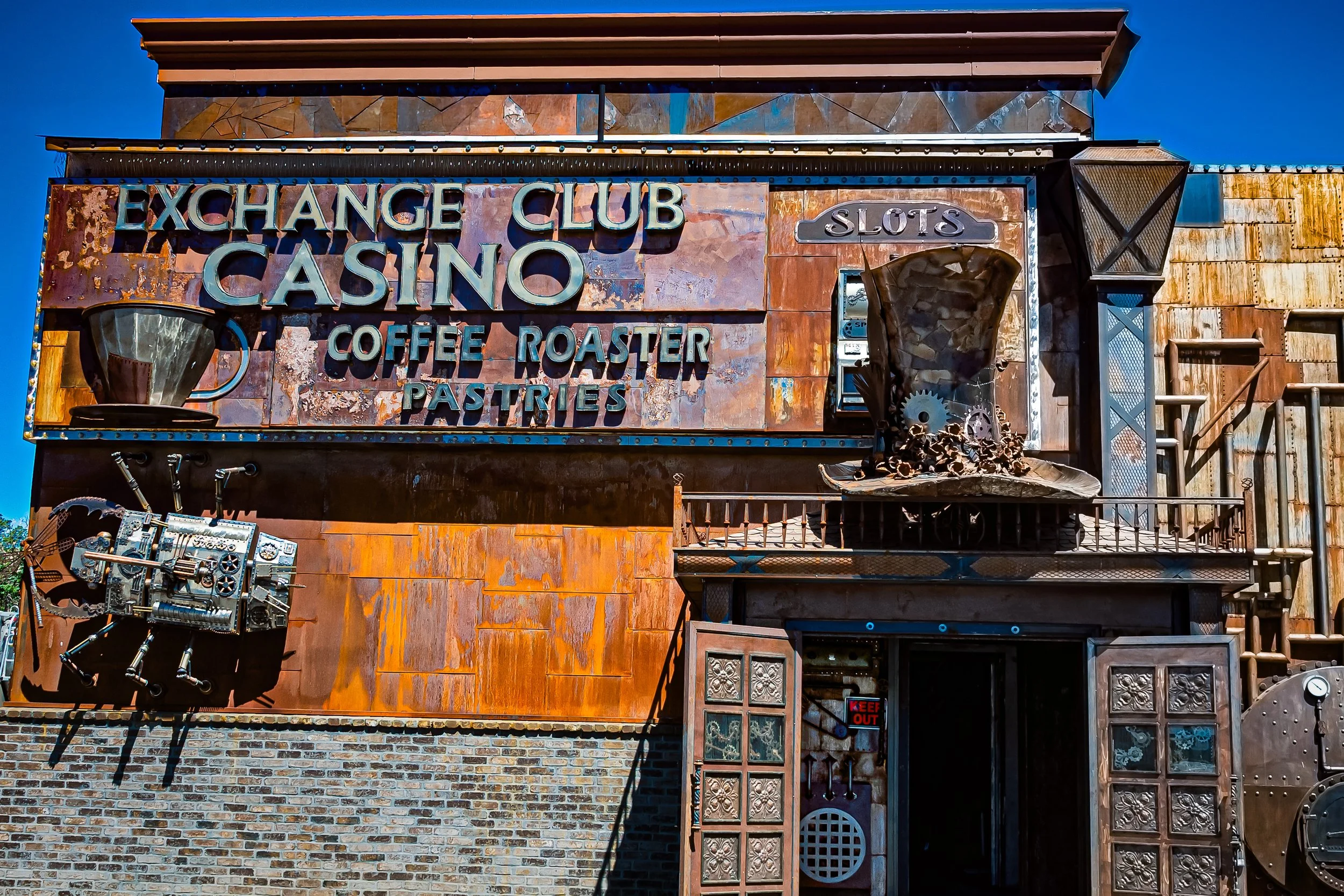 A rusty worn dream stands humming in the street, gears whispering stories., In that cup coffee or more likely bourboun? Beckoning chance, or loss in that heat. Top hat of metal, a robot at the door. Steampunk haven where fortune and steam both roar. 