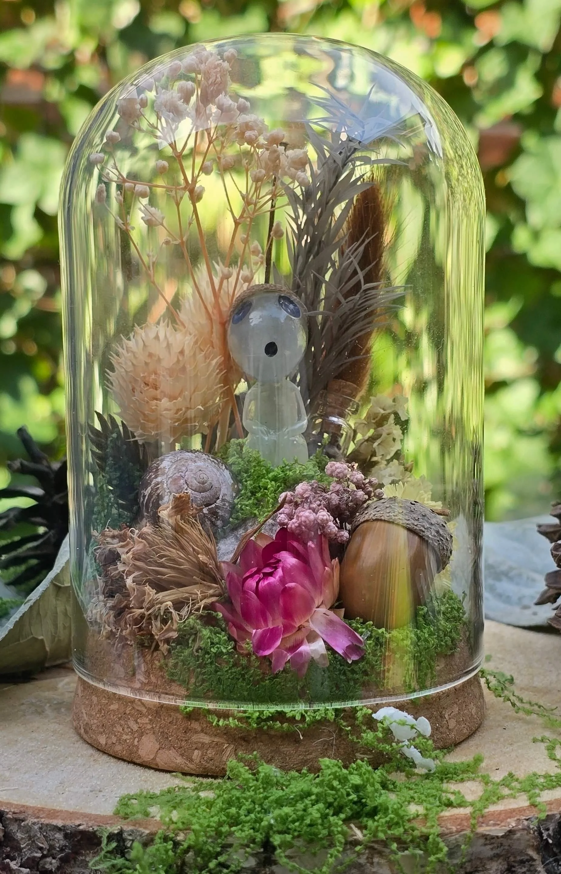 A glass dome containing dried flowers, leaves, shells, a pink lotus flower, and a small figure, set on a wooden base with moss and greenery, outdoors with blurred green foliage in the background.