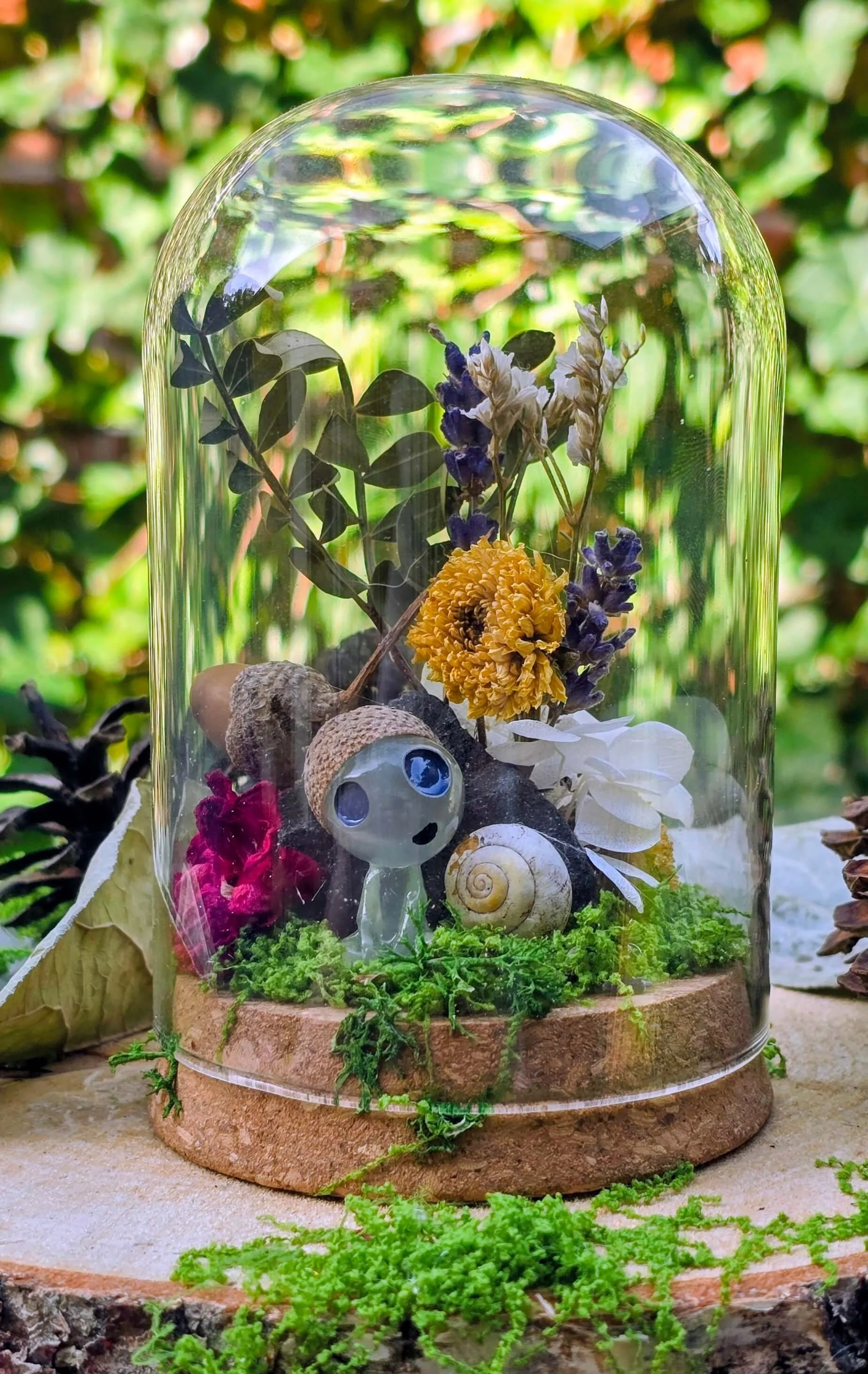 Glass jar with decorative items including a small robot figurine, seashells, dried flowers, and green moss with a leafy background.