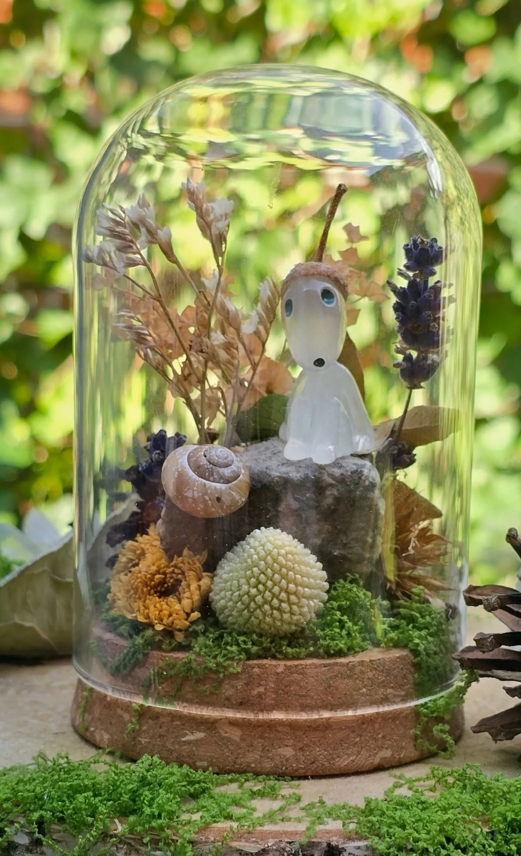 A miniature scene inside a glass terrarium, featuring dried plants, pinecones, moss, and a small white figurine with blue eyes, set against a blurred green outdoor background.