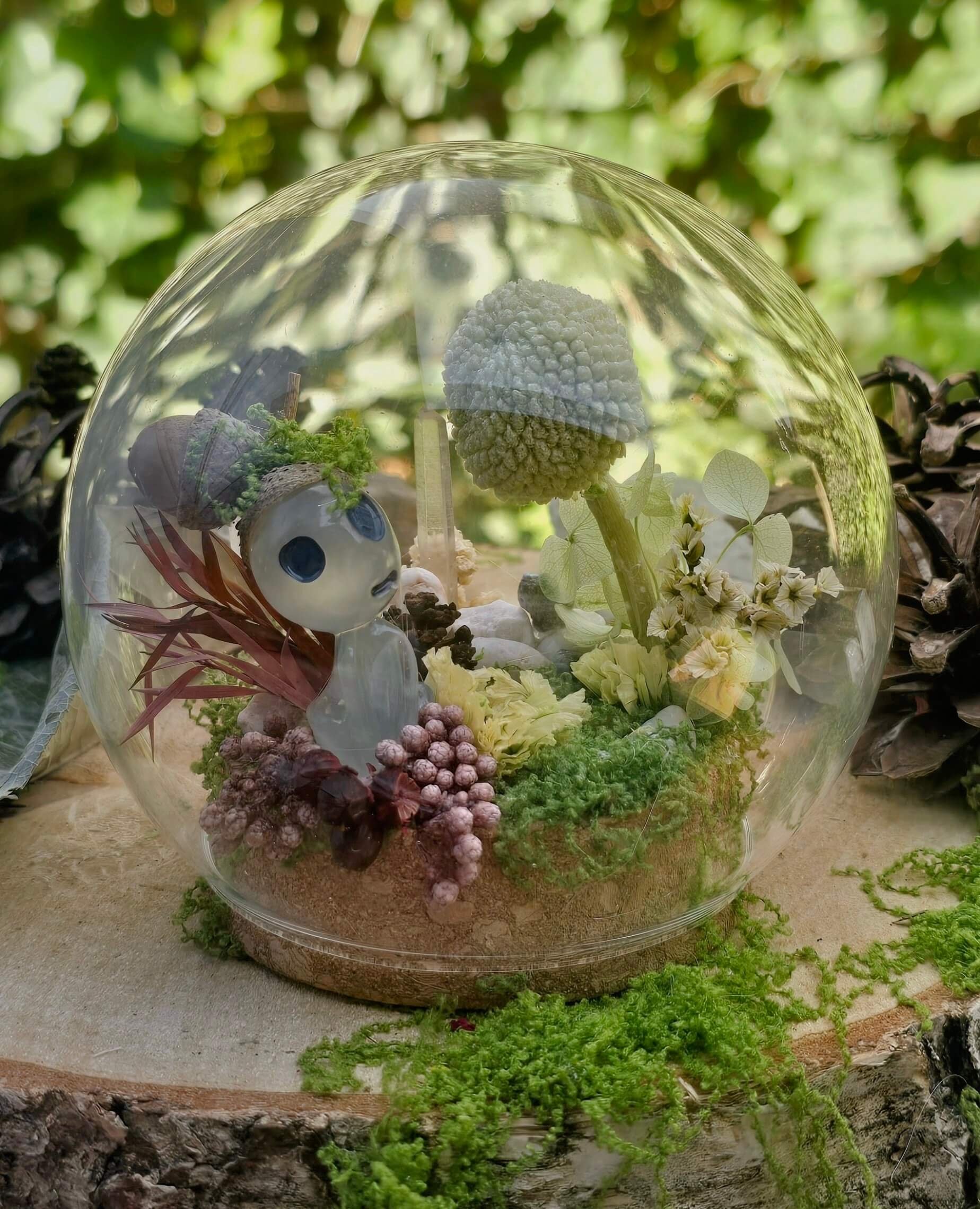 A glass terrarium with various small plants, a skull, and decorative elements, placed on a surface outdoors with green foliage in the background.