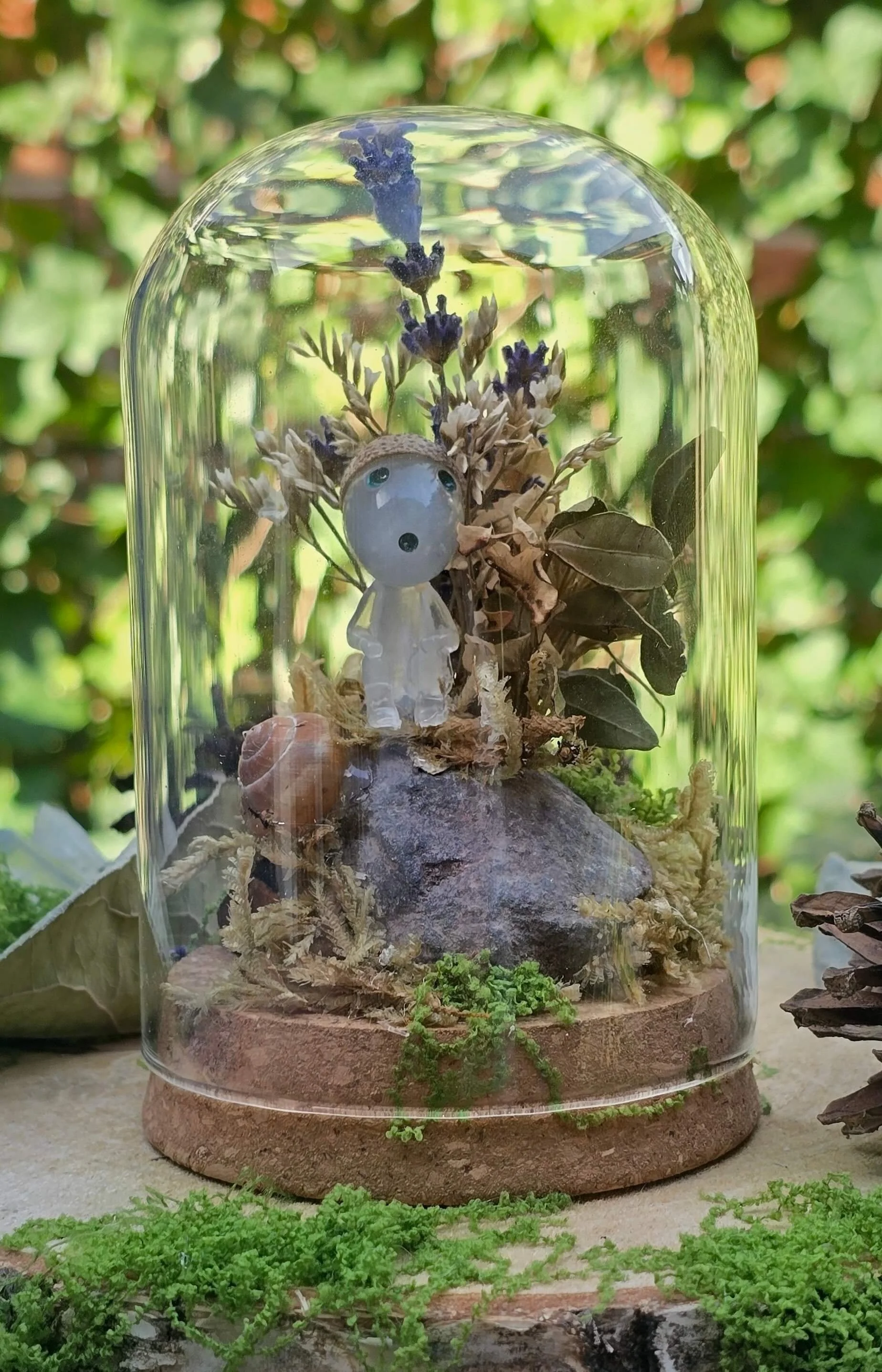A glass terrarium with dried flowers, a small figurine resembling a person, and small green plants inside, set on a round clay base. The background is green foliage.