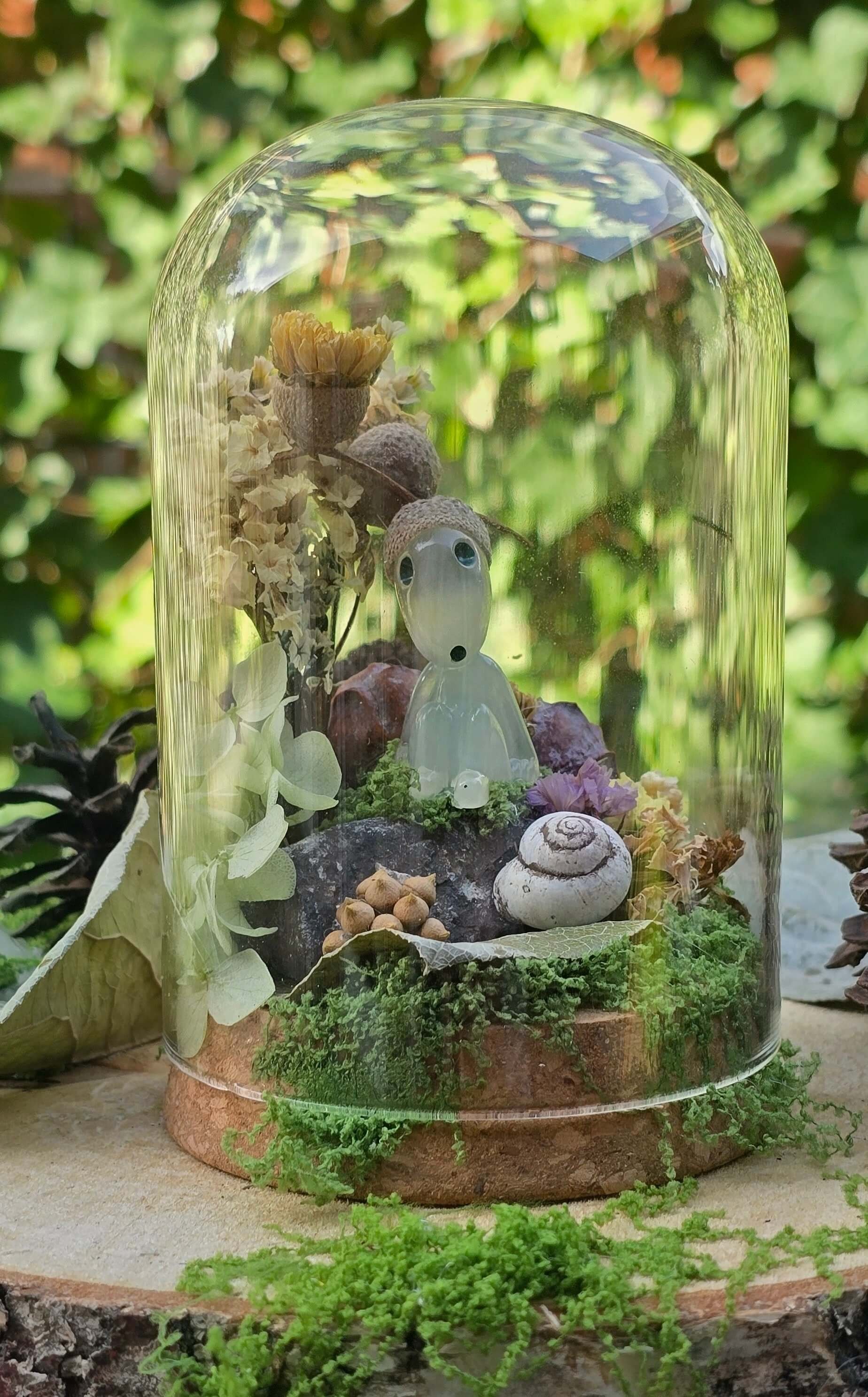 A miniature fairy garden display inside a glass dome featuring small plants, shells, and figurines, with green foliage background.