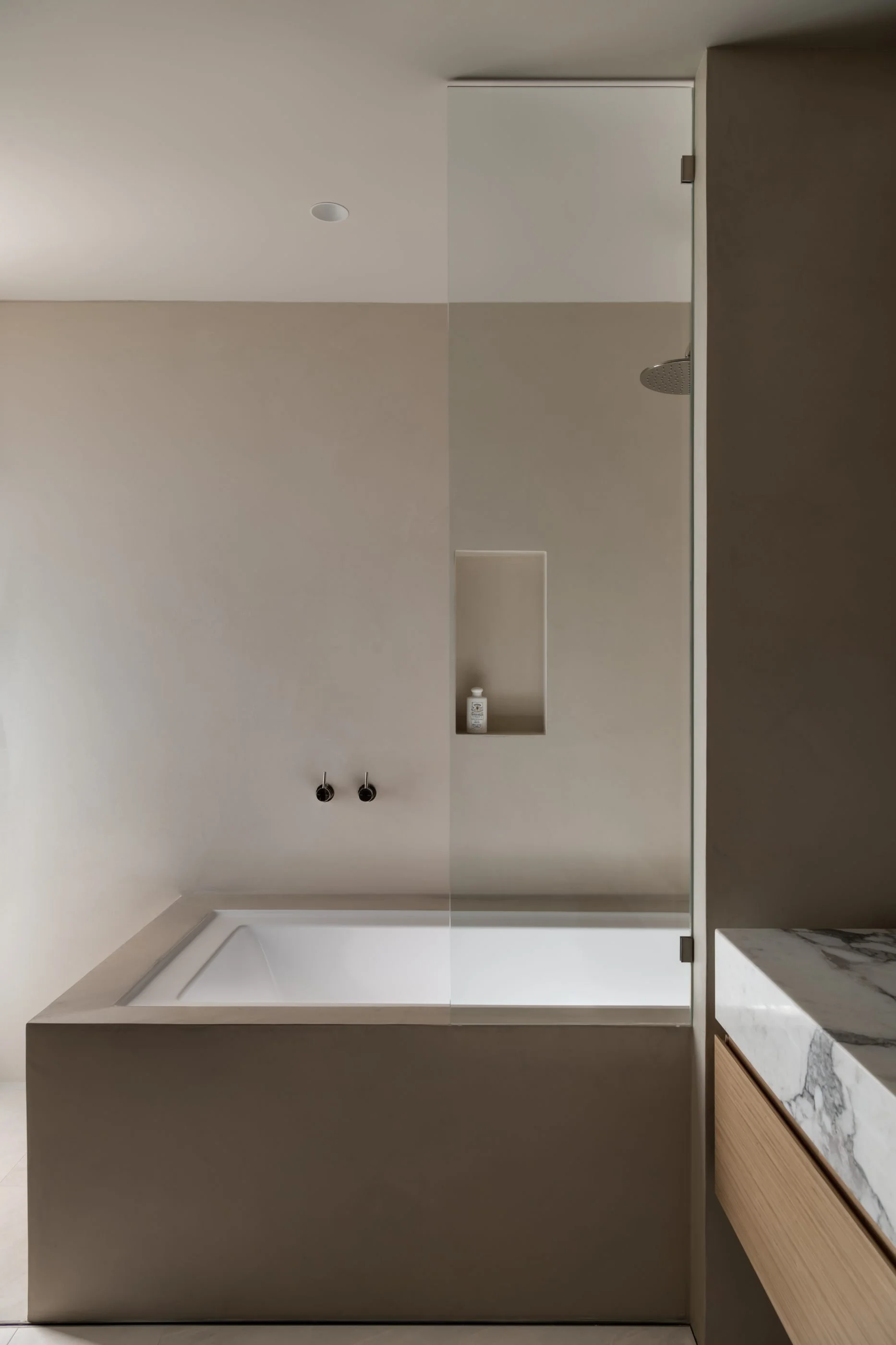 A modern bathroom with a bathtub, wall-mounted showerhead, a built-in shower niche with a bottle inside, and a marble countertop with wooden cabinetry.