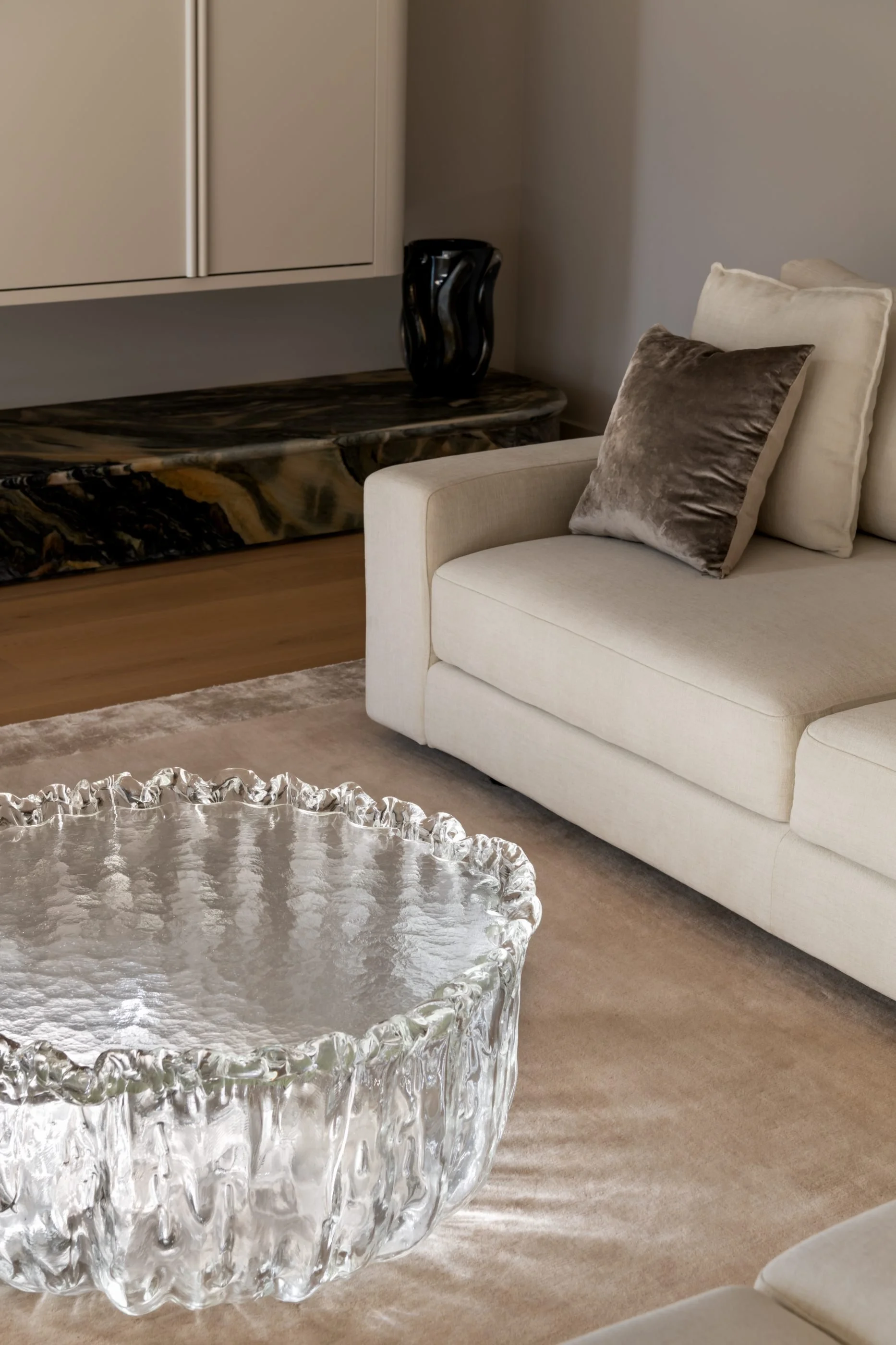 A living room with a white sofa and dark velvet pillows, a marble console table with a black decorative vase, a glass coffee table, and a beige carpet.