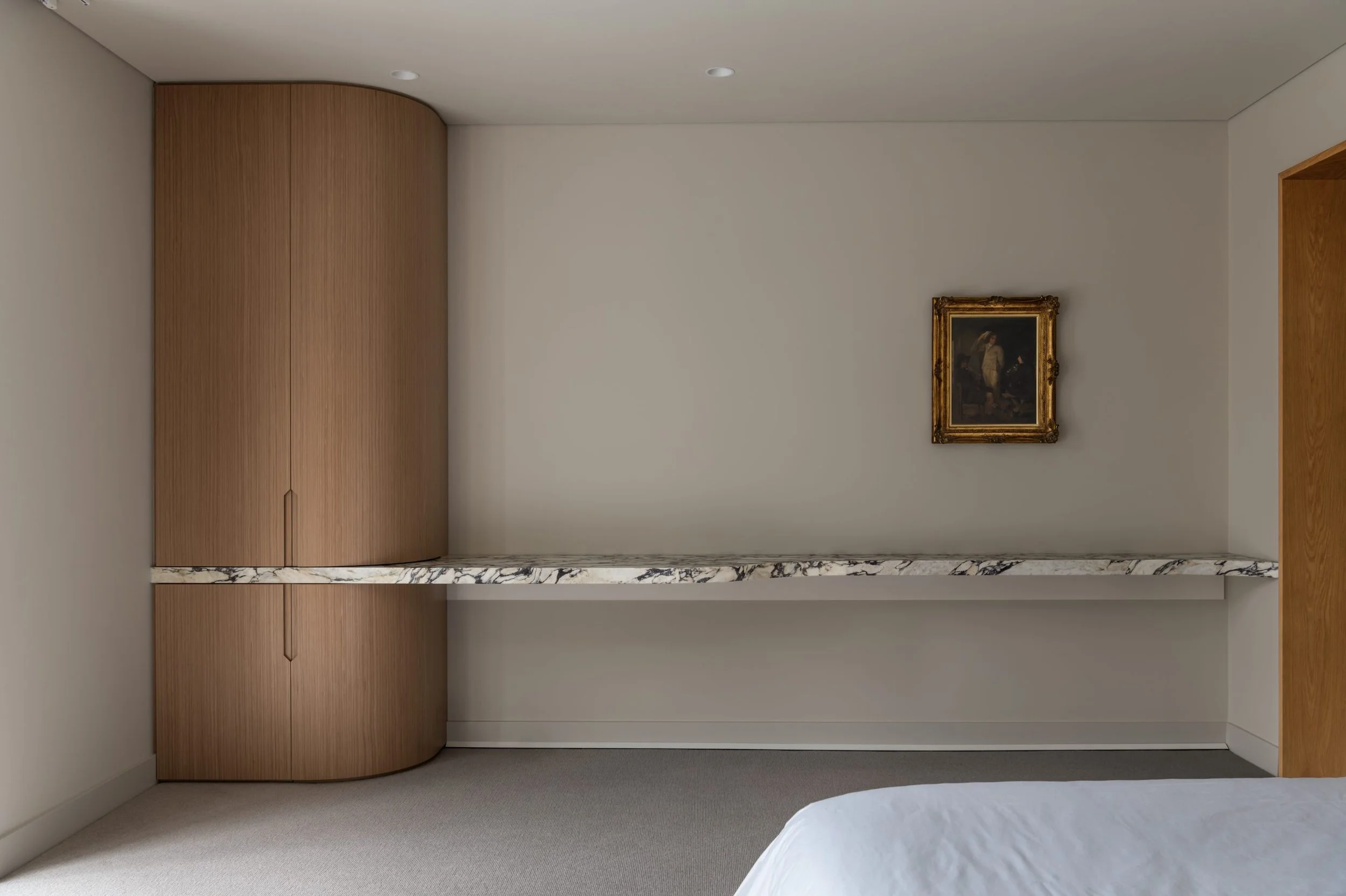 A minimalist hotel room interior with a beige wall, a framed painting, a marble shelf, wooden panel, and part of a bed with white sheets visible.