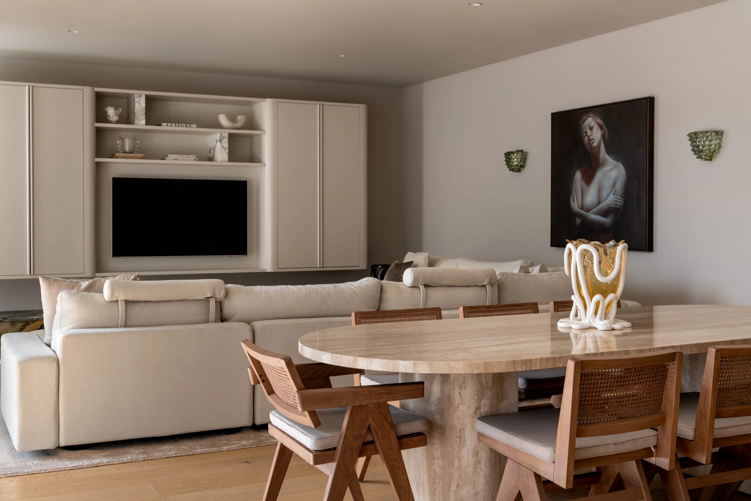 A living room with a cream-colored sofa, a wooden dining table with chairs, a wall-mounted television, white built-in shelves with decorative items, a large portrait painting of a woman, and a decorative ceramic vase on the table.