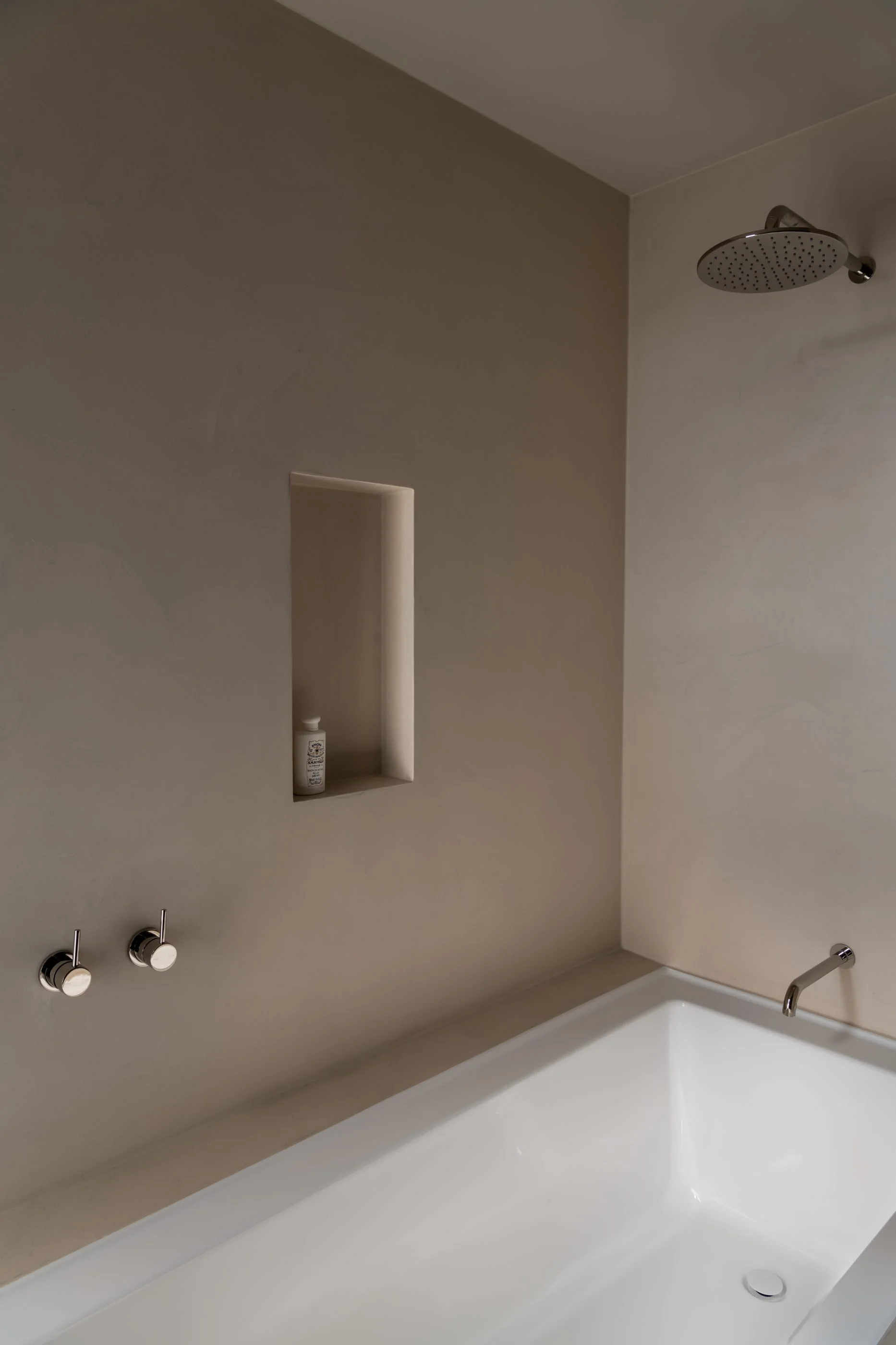 Bathroom with a white bathtub, a showerhead attached to the ceiling, two silver faucets, and a built-in wall niche with a soap bottle inside.