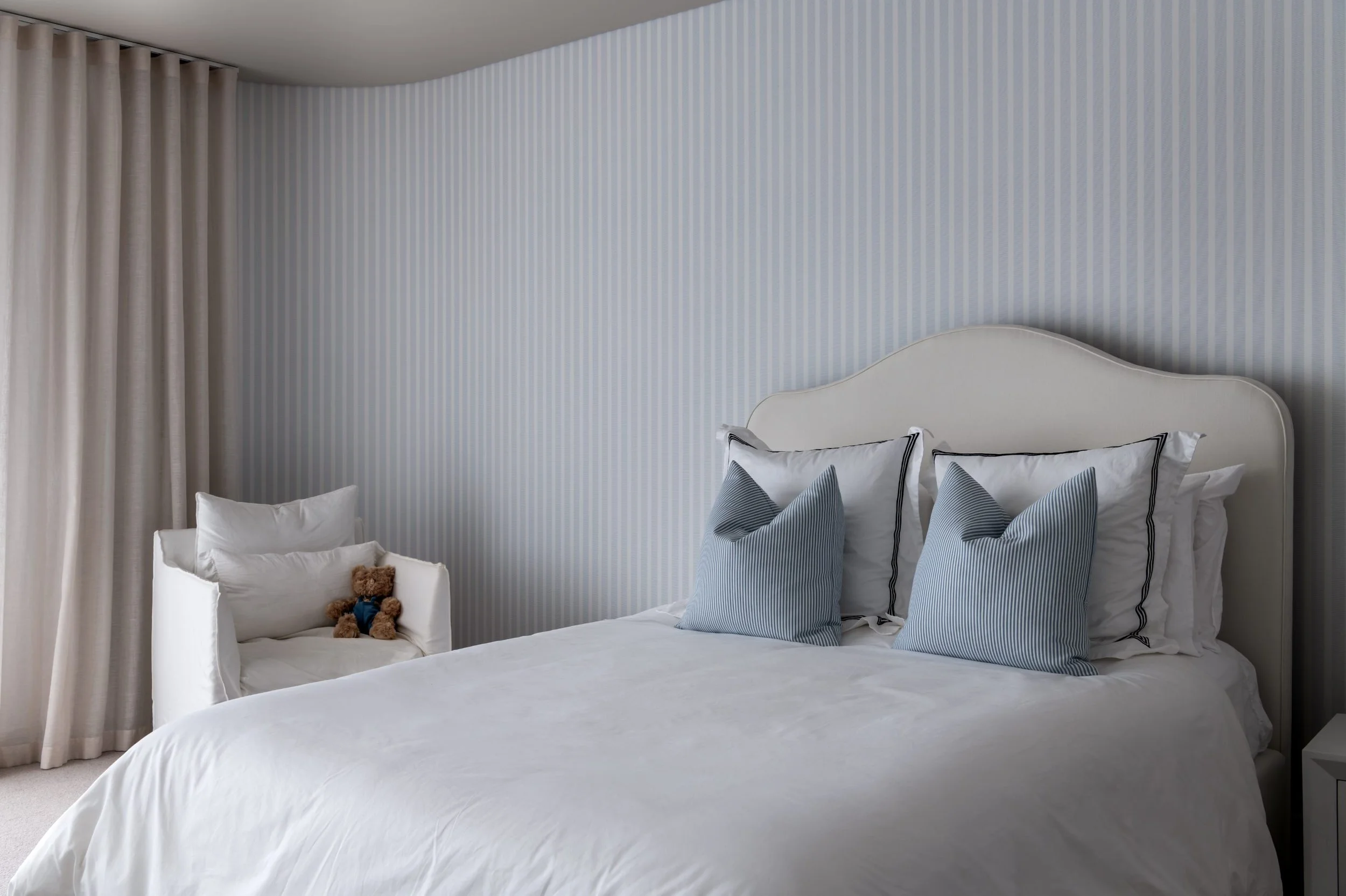 A neatly made bed with white bedding and pillows, positioned against a striped blue and white wallpapered wall, with a white headboard. To the left, there is a white chair with a stuffed teddy bear on it, near beige curtains.