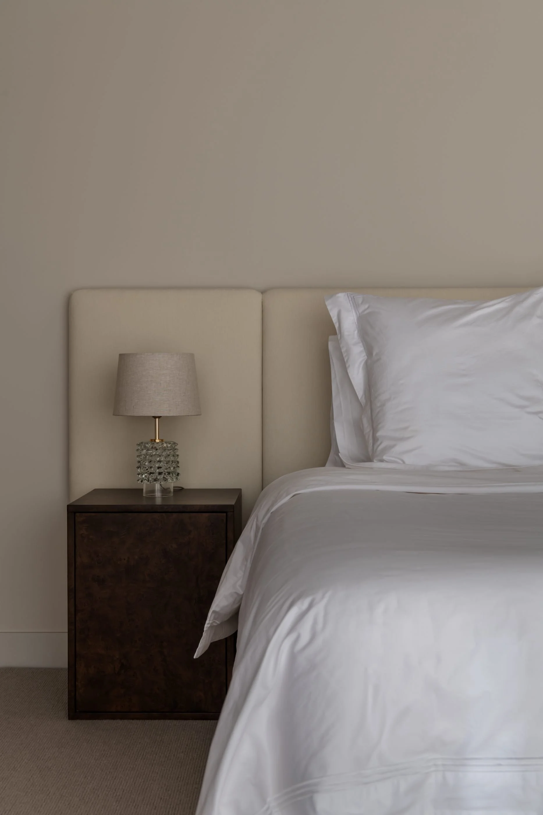 A bedroom with a beige upholstered headboard, a nightstand with a textured glass lamp, and white bedding with a pillow on a beige carpeted floor.
