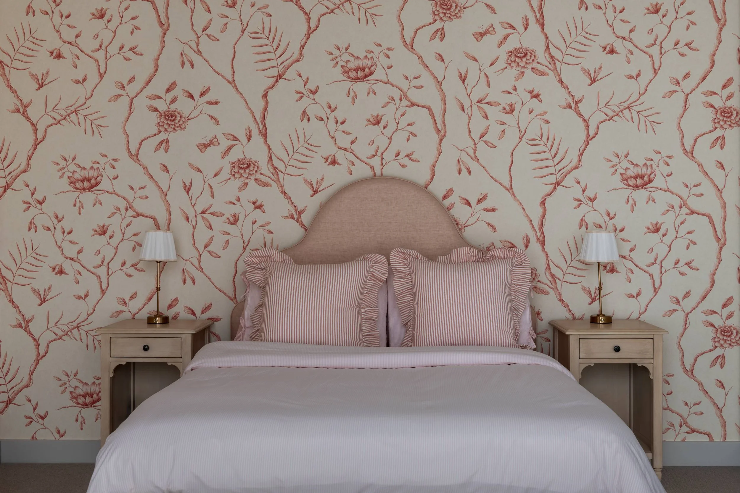 A bedroom with a pink floral wallpaper background behind a bed with ruffled pink and white striped pillows and a matching bedspread. There are two white nightstands on either side of the bed, each with a white lamp with a gold base. The headboard is 