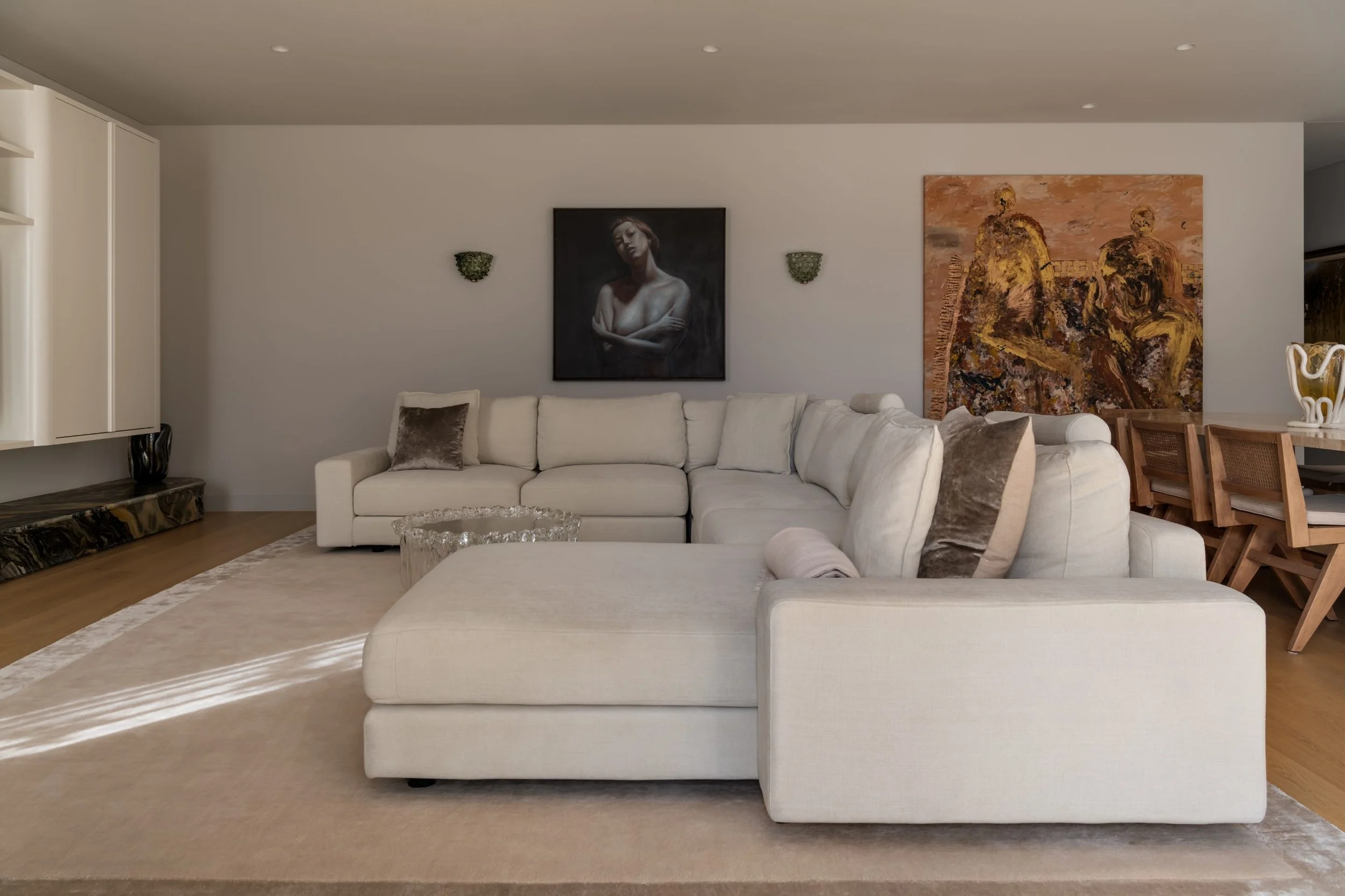 Living room with a large white sectional sofa, wall art including a portrait of a woman and an abstract painting, a glass coffee table, and a dining area with wooden chairs and a white decorative vase.