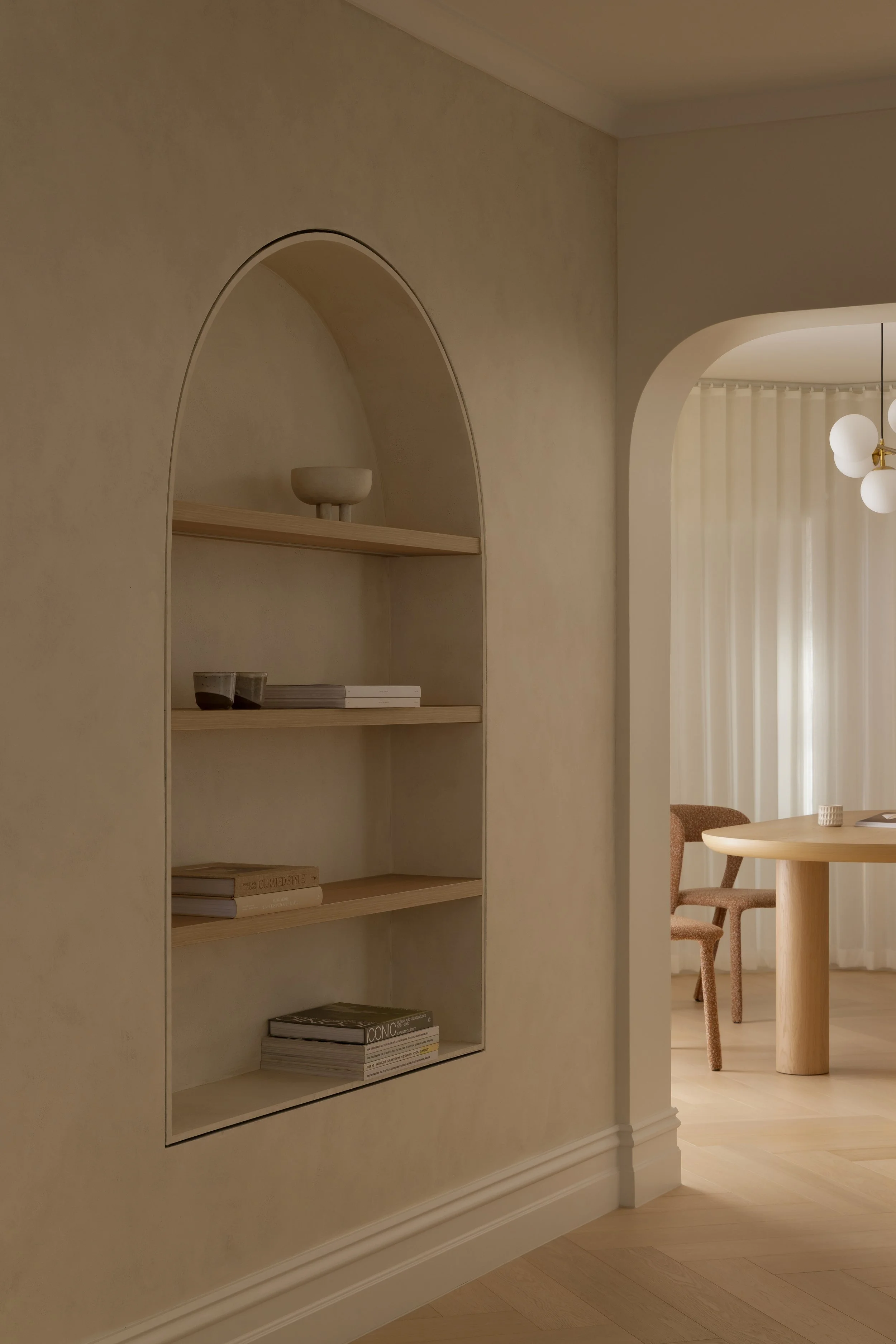 A minimalist room with built-in shelves in an alcove on a beige wall, some books, cups, and decorative objects, next to a dining area with a round wooden table and pink upholstered chairs, in a space with a modern chandelier and cream curtains.
