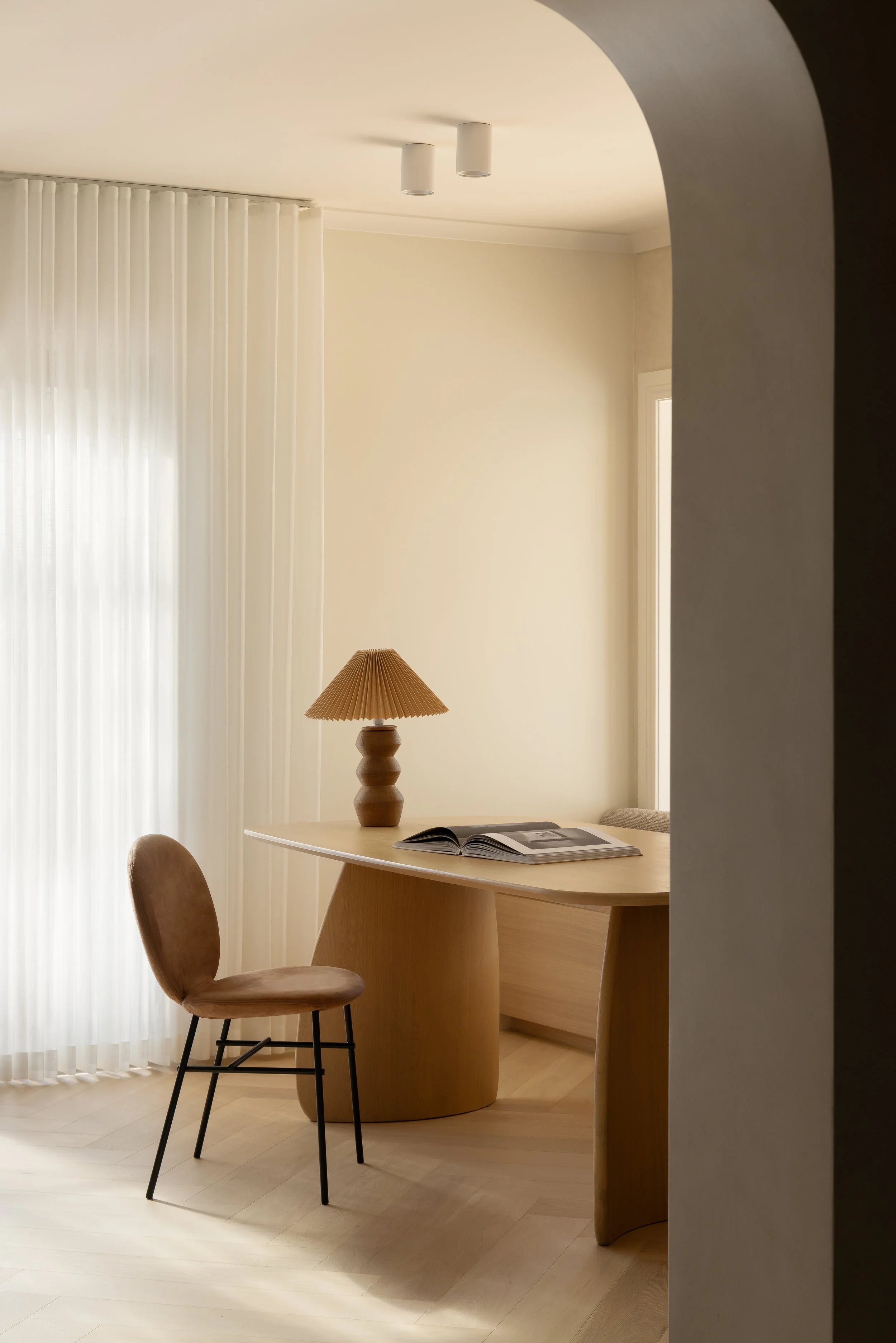 A cozy corner of a room featuring a wooden table with a book open on it, a pinkish-brown upholstered chair, a table lamp with a pleated shade, and sheer white curtains allowing natural light to fill the space.