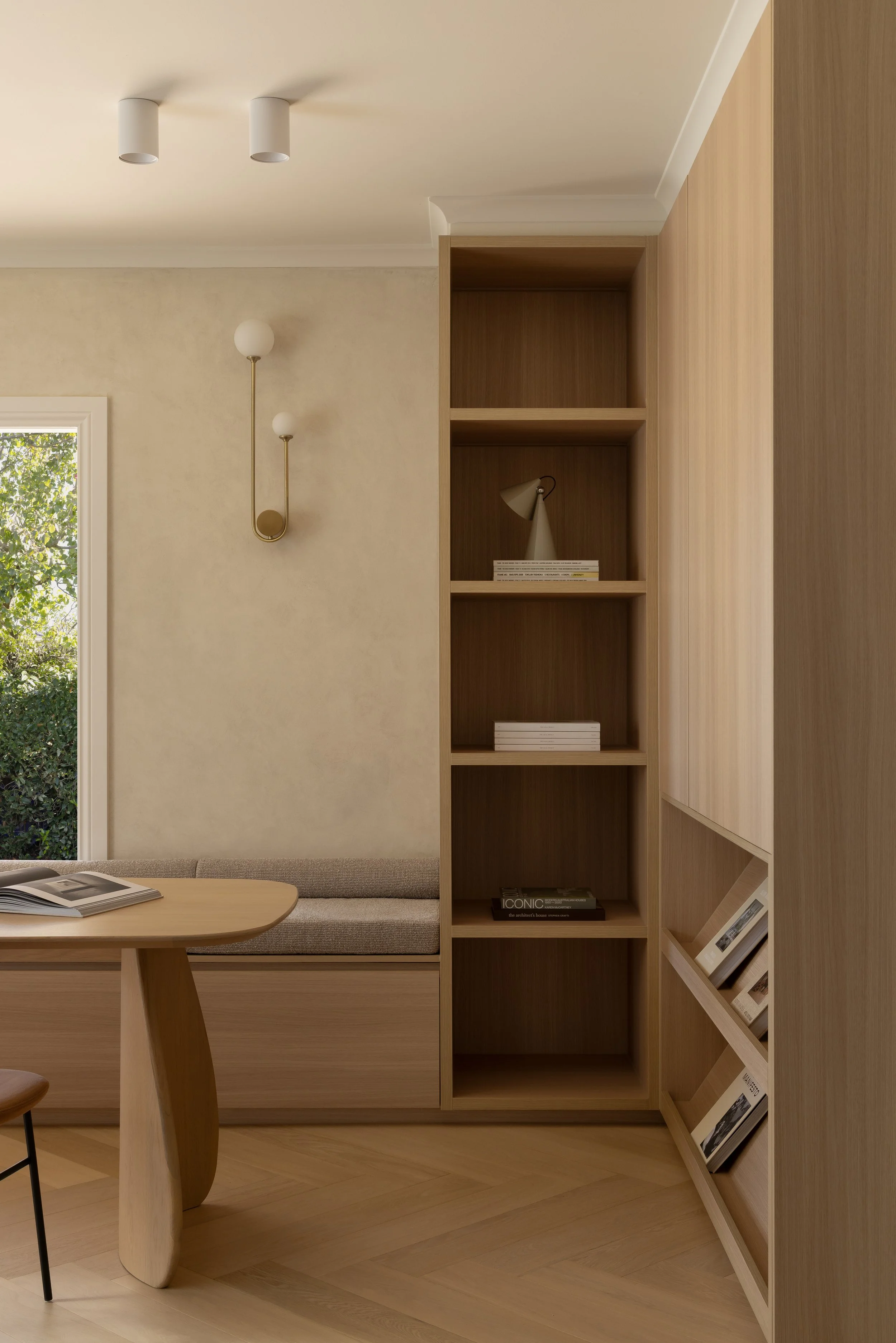 Interior of a modern room with wooden built-in shelves, a small wooden table, and a cushioned window seat. The room has beige walls, a ceiling with white cylindrical spotlights, and a brass wall sconce. Green foliage is visible outside the window.