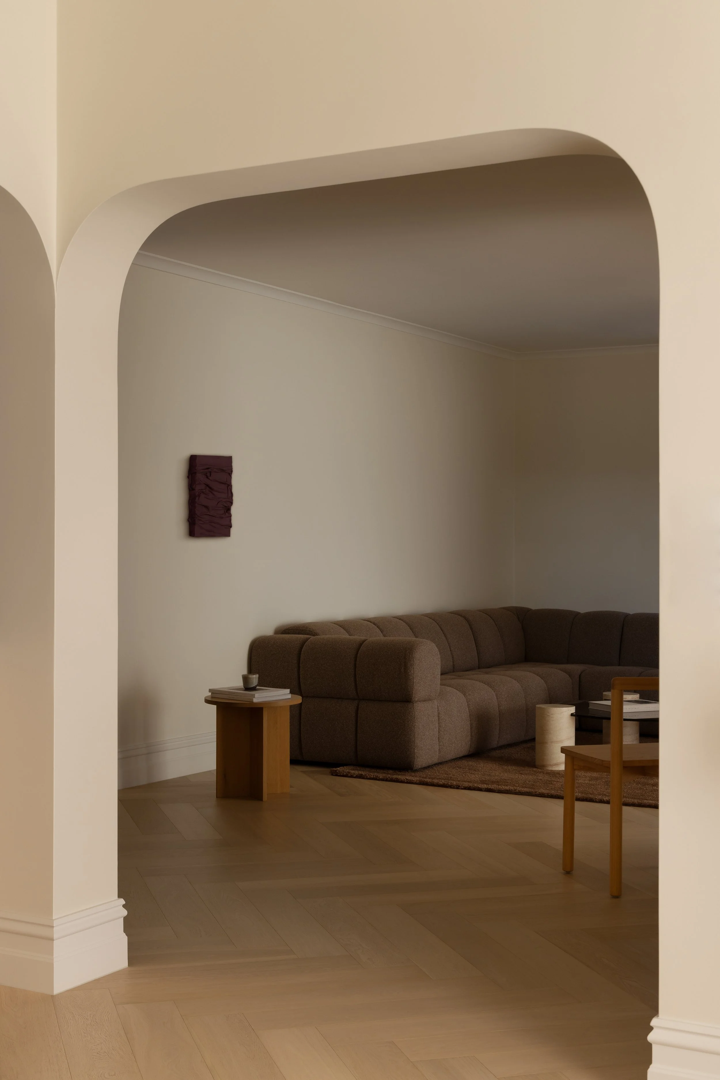 A cozy living room with beige walls, a brown sectional sofa, a small wooden side table with books and a candle, and modern decor including a mauve wall art piece.