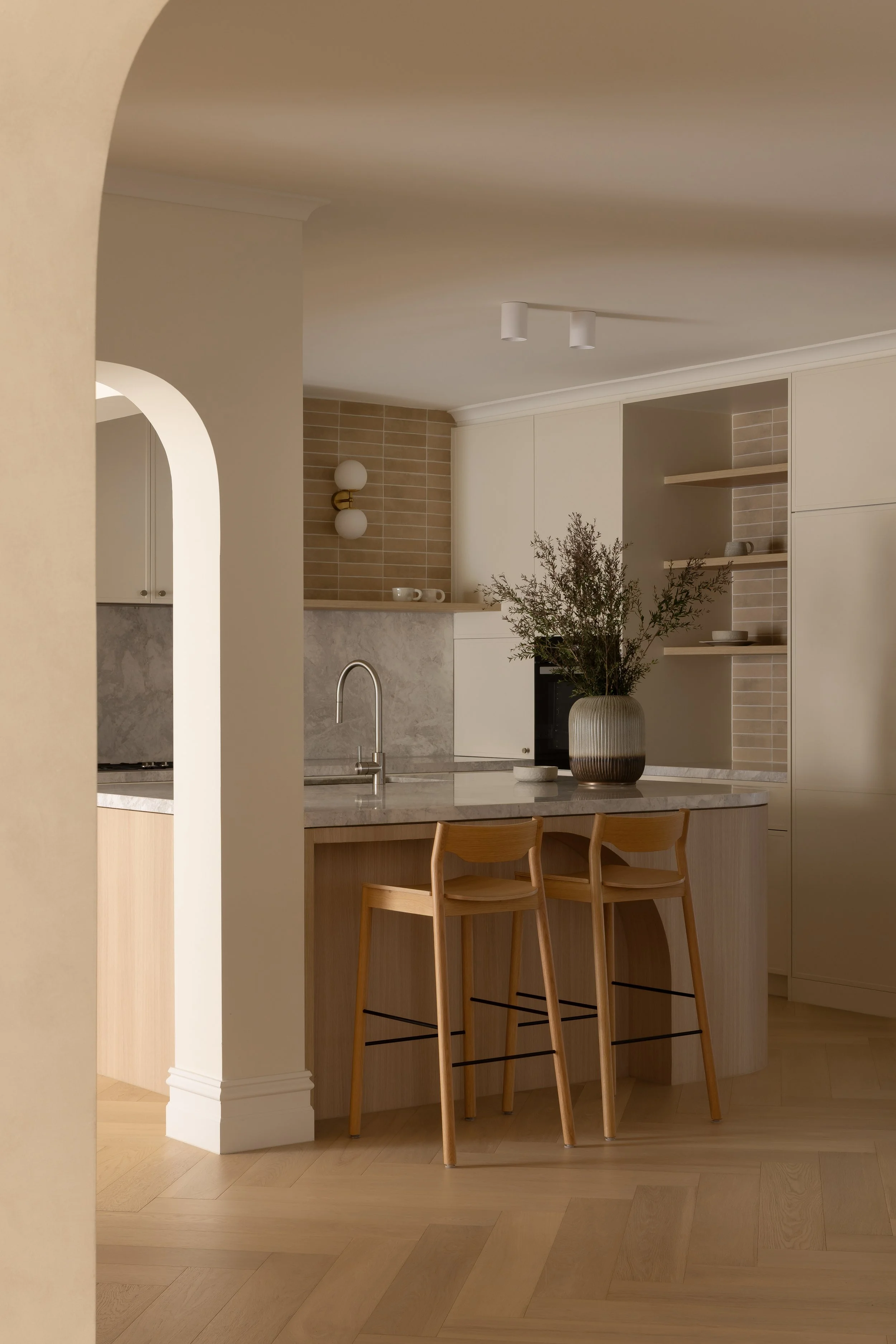 Modern kitchen with light wood cabinets, a marble island, and wooden bar stools, decorated with a potted plant and minimalist wall lights.