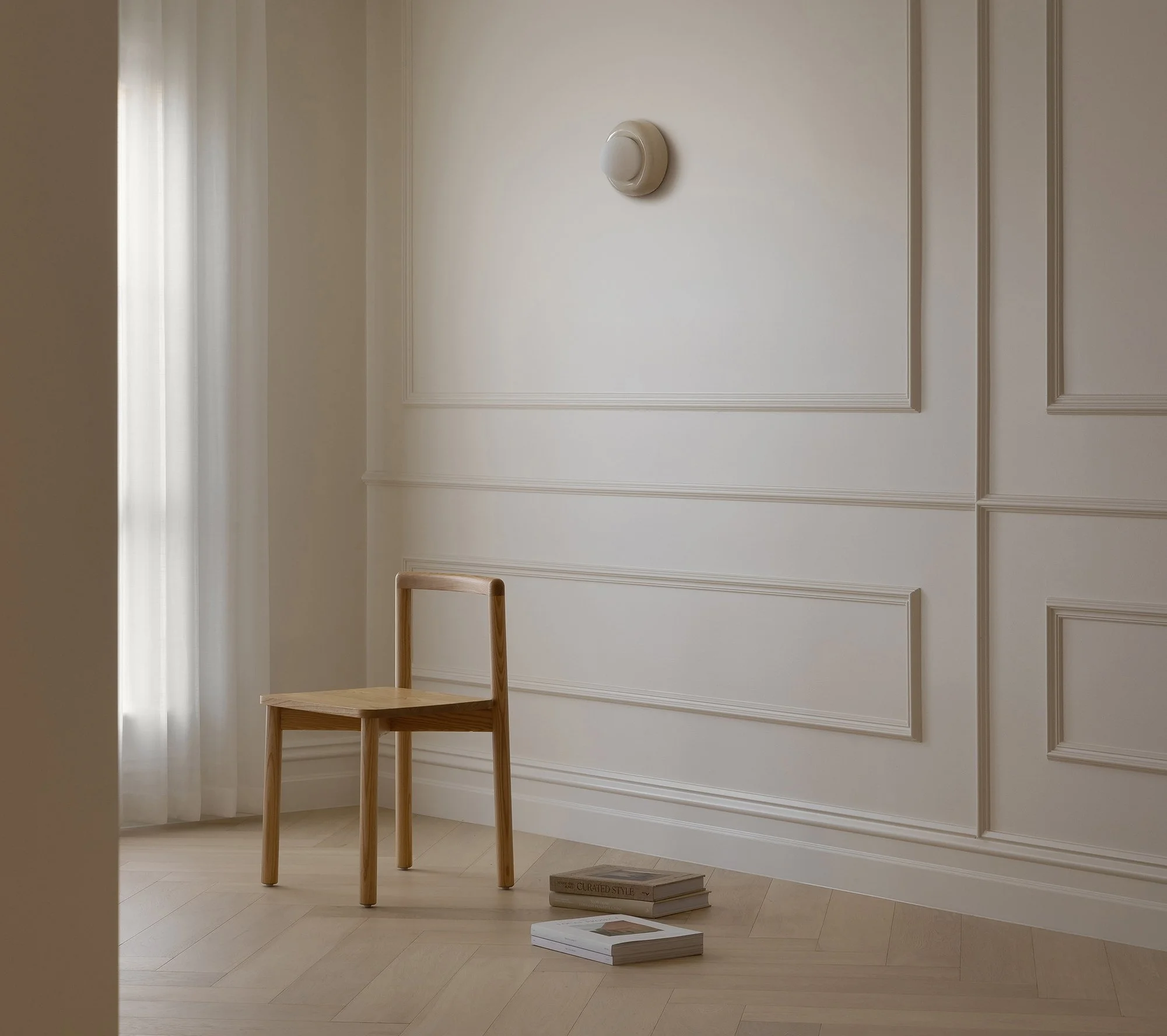 A single wooden chair with no cushions sits in an empty room with white walls and wooden flooring. The wall has decorative molding, and there are a few books on the floor near the chair, with one that reads "Curated Style." Natural light filters through sheer curtains on a window to the left.