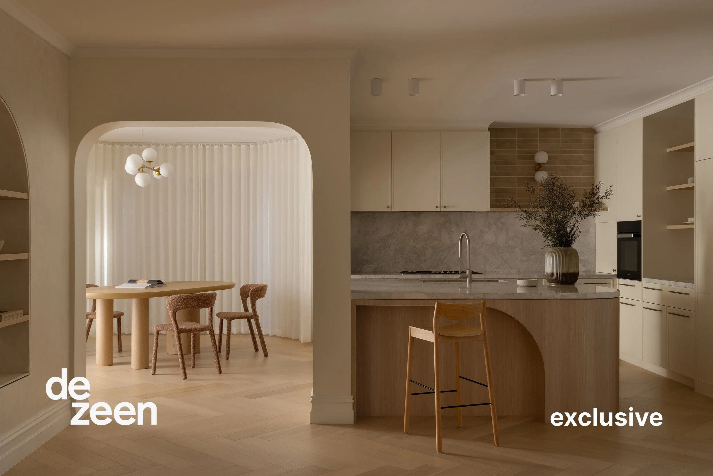 Interior view of a modern kitchen with wooden cabinets, a marble countertop, and a plant in a large vase. Adjacent dining area with a round wooden table, four chairs, and a ceiling light fixture with multiple globes. Light-colored flooring and soft lighting create a cozy atmosphere.
