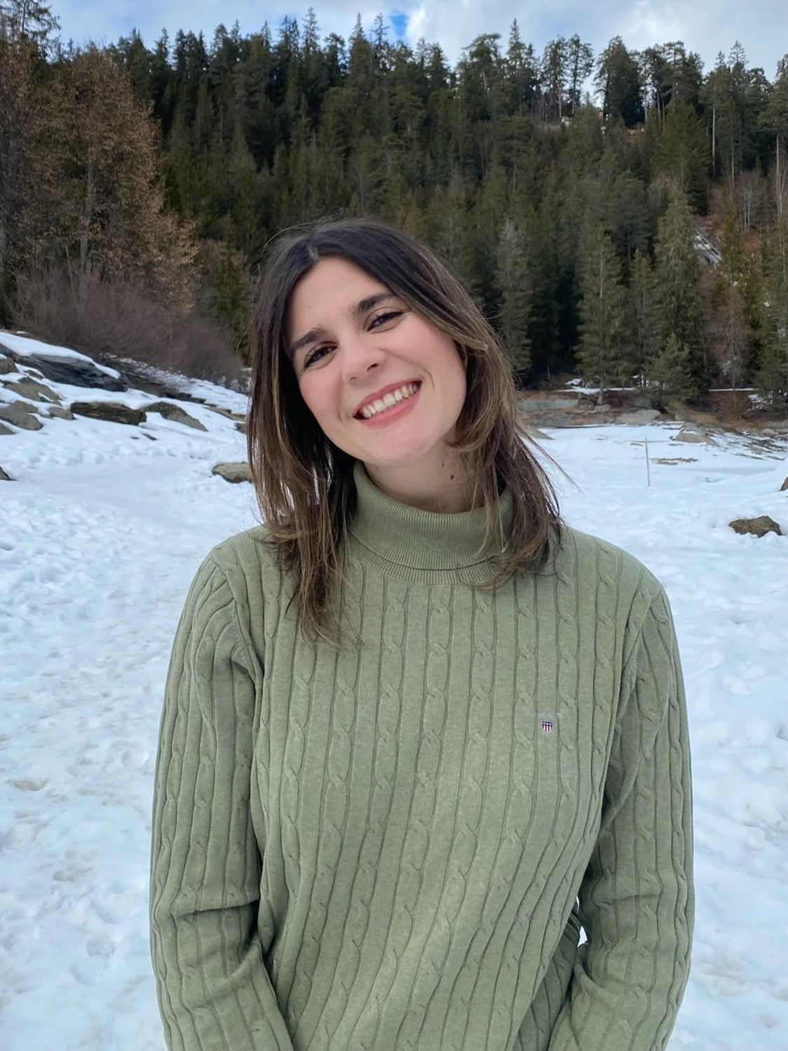 Young woman with shoulder-length dark hair smiling outdoors in a snowy landscape with trees in the background.