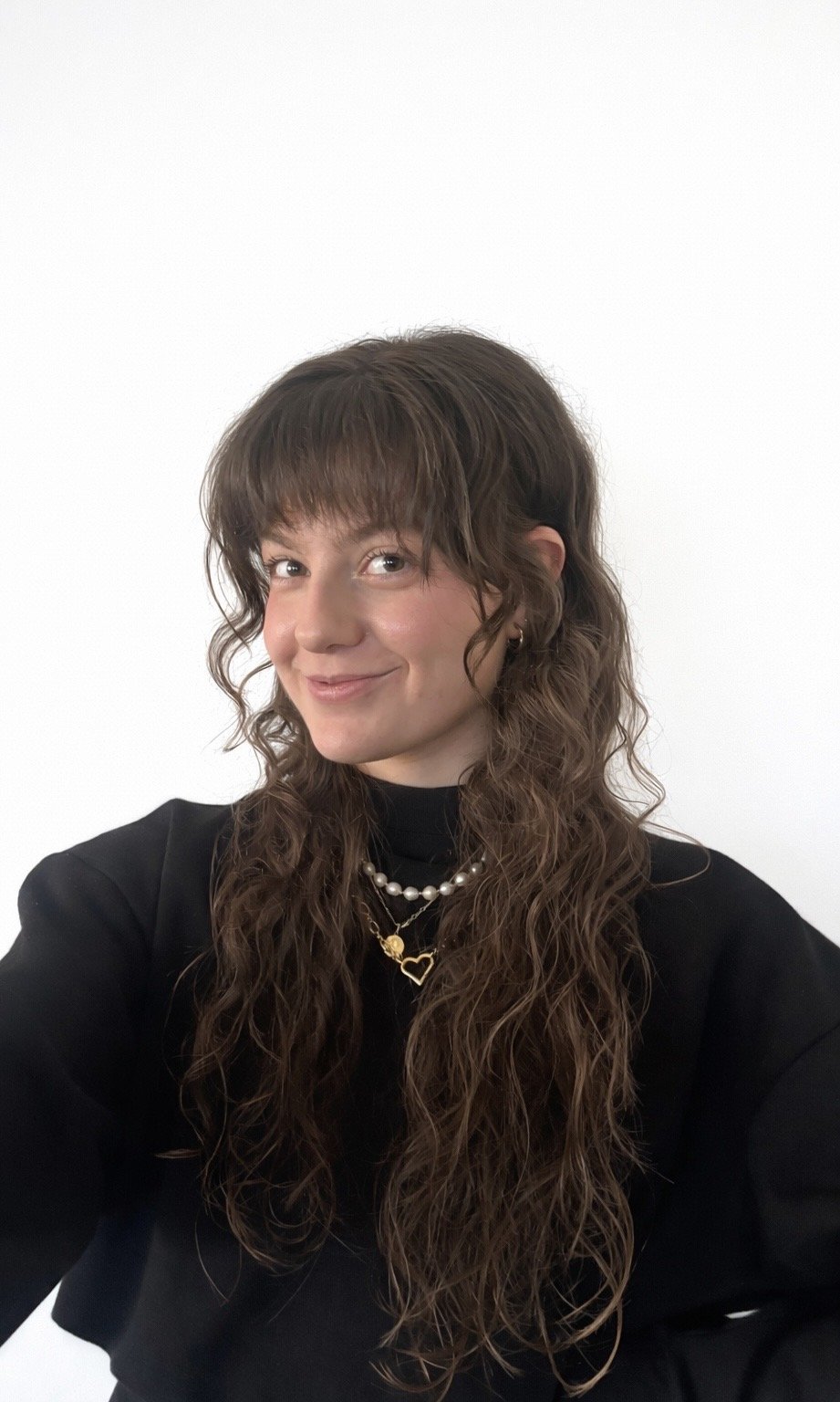 Young woman with long curly brown hair wearing a black top, gold heart necklace, pearl necklace, and earrings, smiling against a plain white background.