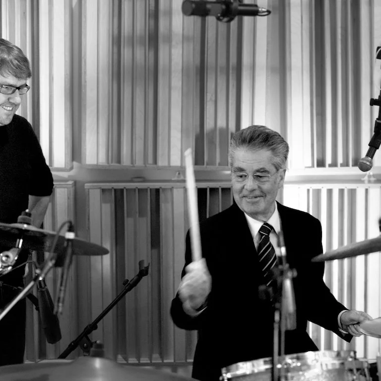 An older man in a suit playing the drums in a recording studio, with a younger man smiling nearby.
