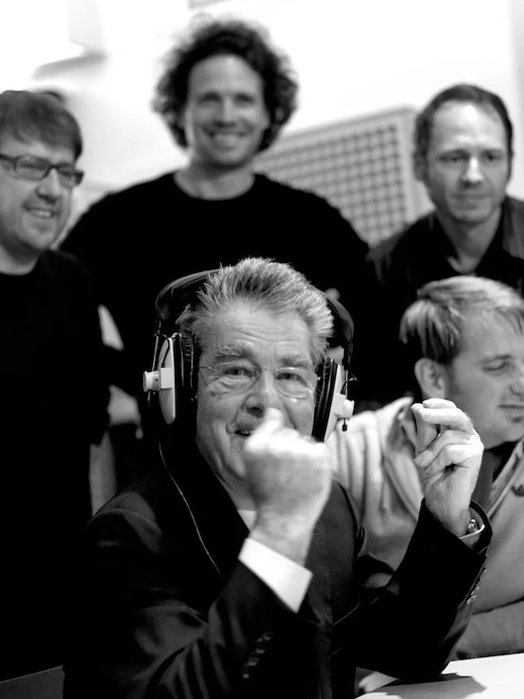 A group of five men gathered around a table, with one man wearing large headphones, smiling and making a fist gesture.