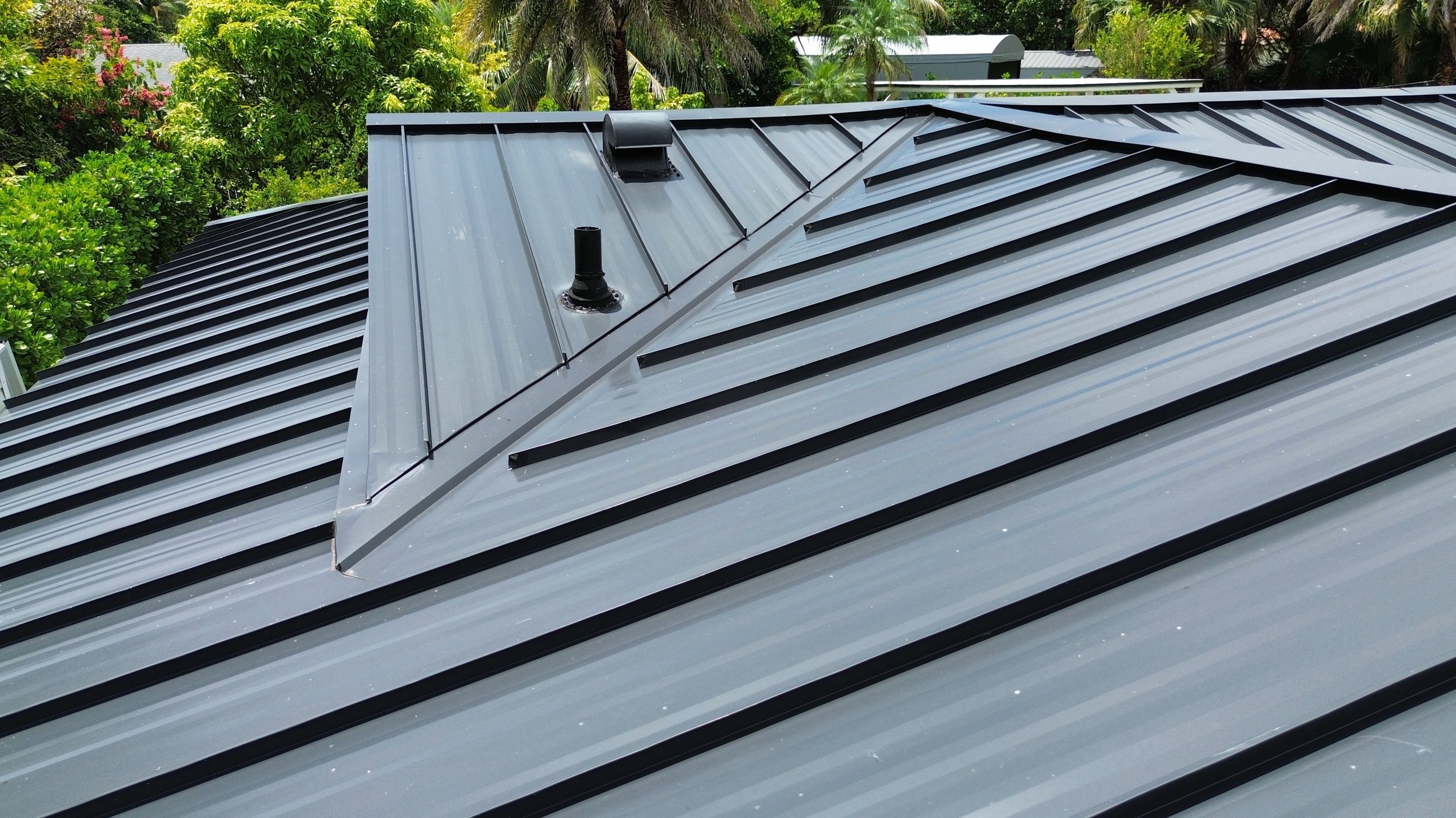 Metal roof with vents and a 'V' shaped ridge, surrounded by green trees and sky.