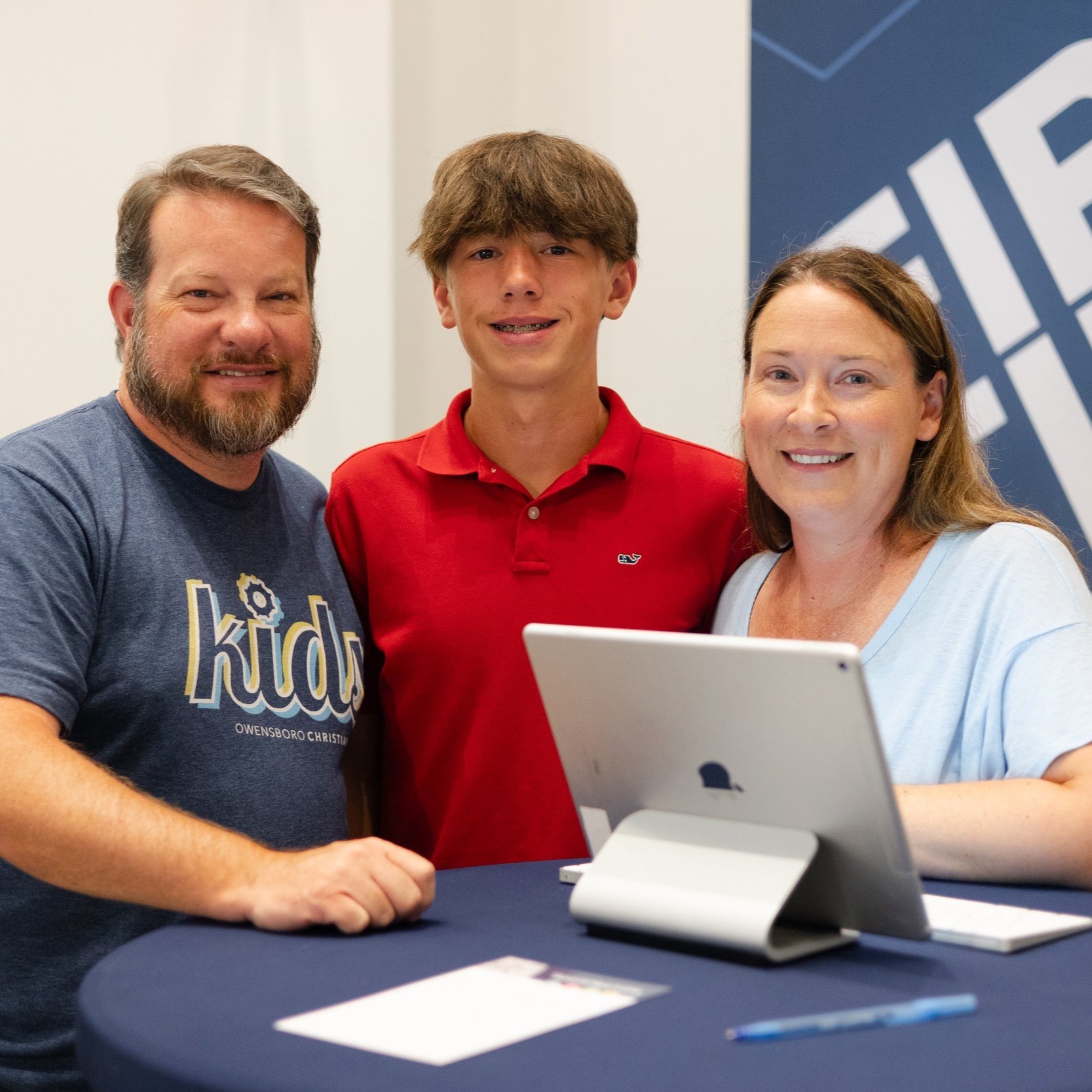 A group of three people, including a young man, a woman, and a man, standing behind a table with a tablet computer, smiling at the camera.