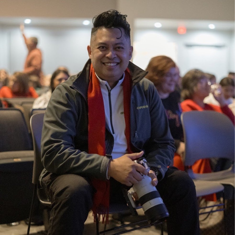 A man smiling and holding a camera, sitting in a room filled with seated people.