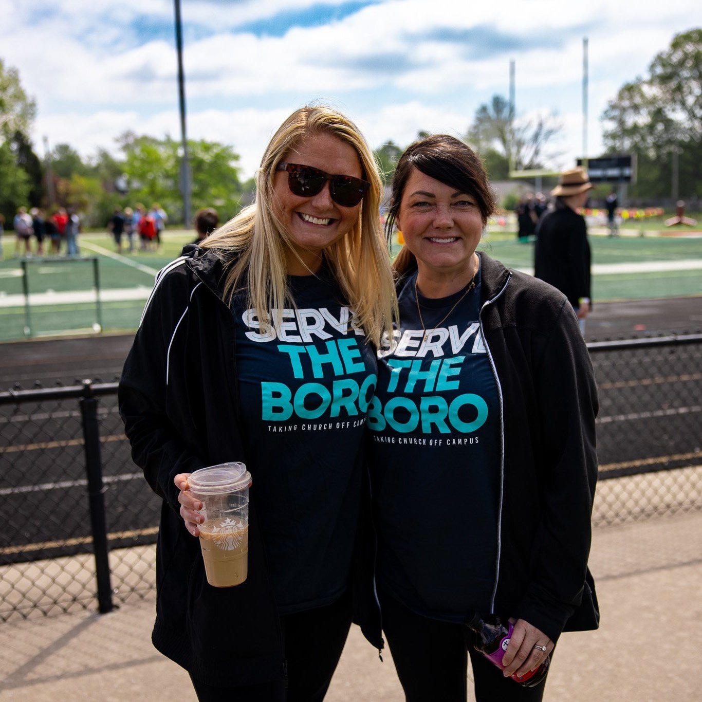 Two women smiling and standing outdoors at a sports field or track, wearing matching black t-shirts with blue and white text, black jackets, and one holding a clear plastic cup with a beverage.