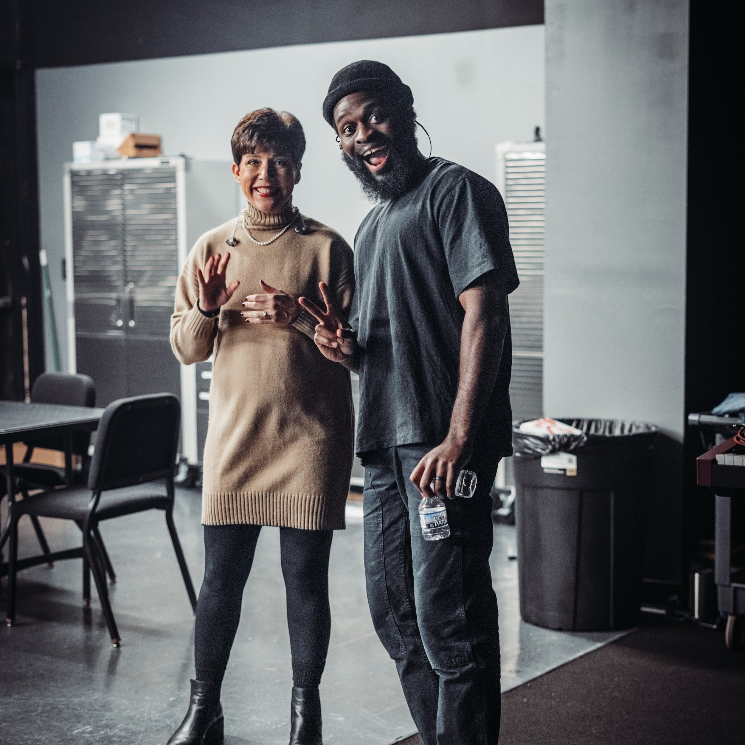 Two people, a woman and a man, standing indoors, smiling, with the woman waving and the man holding a water bottle, in a room with chairs and equipment in the background.