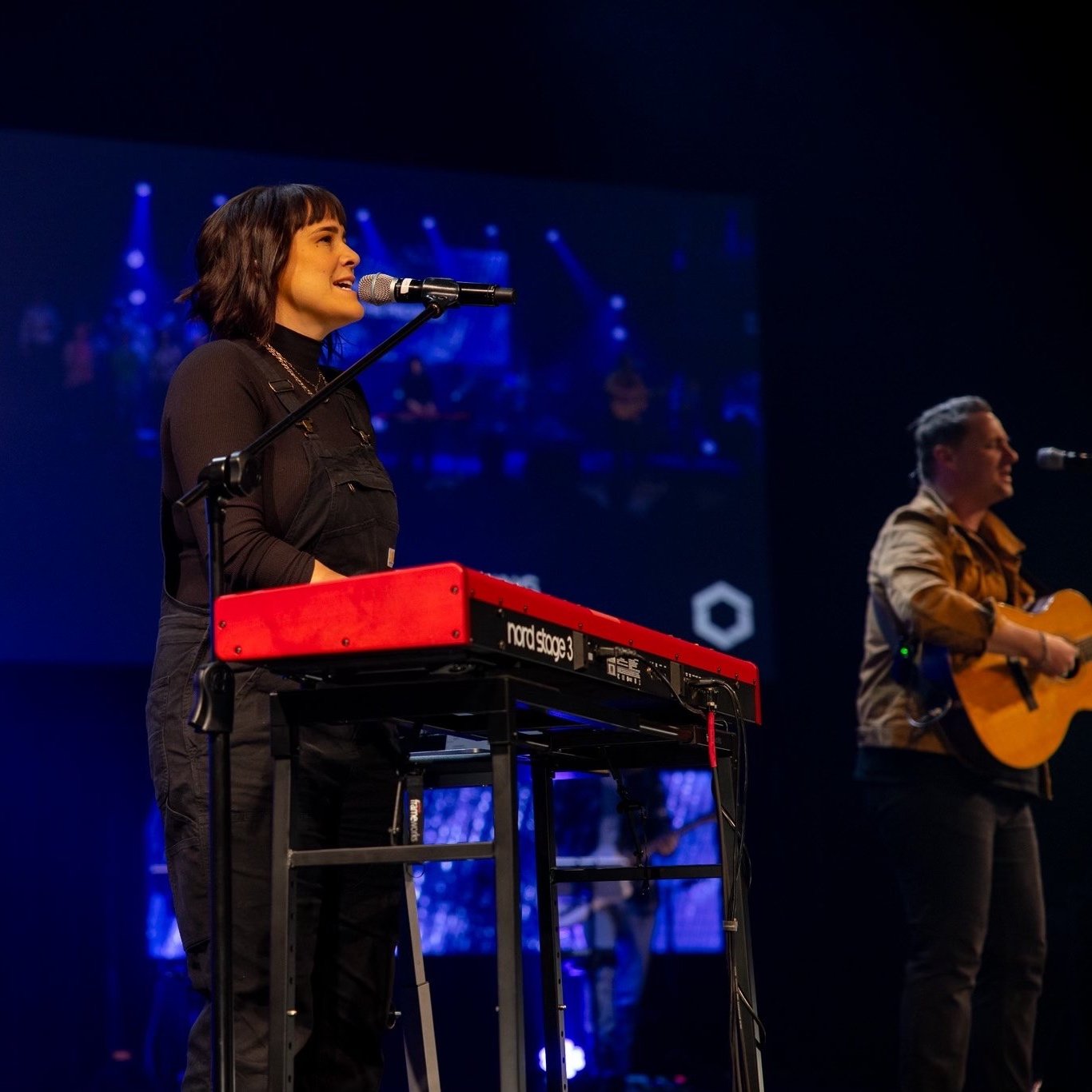 A woman with dark, shoulder-length hair singing into a microphone while playing a red Nord Stage 3 keyboard on stage. A man with short hair plays an acoustic guitar beside her. The background has dark lighting with blue accents and a large screen dis