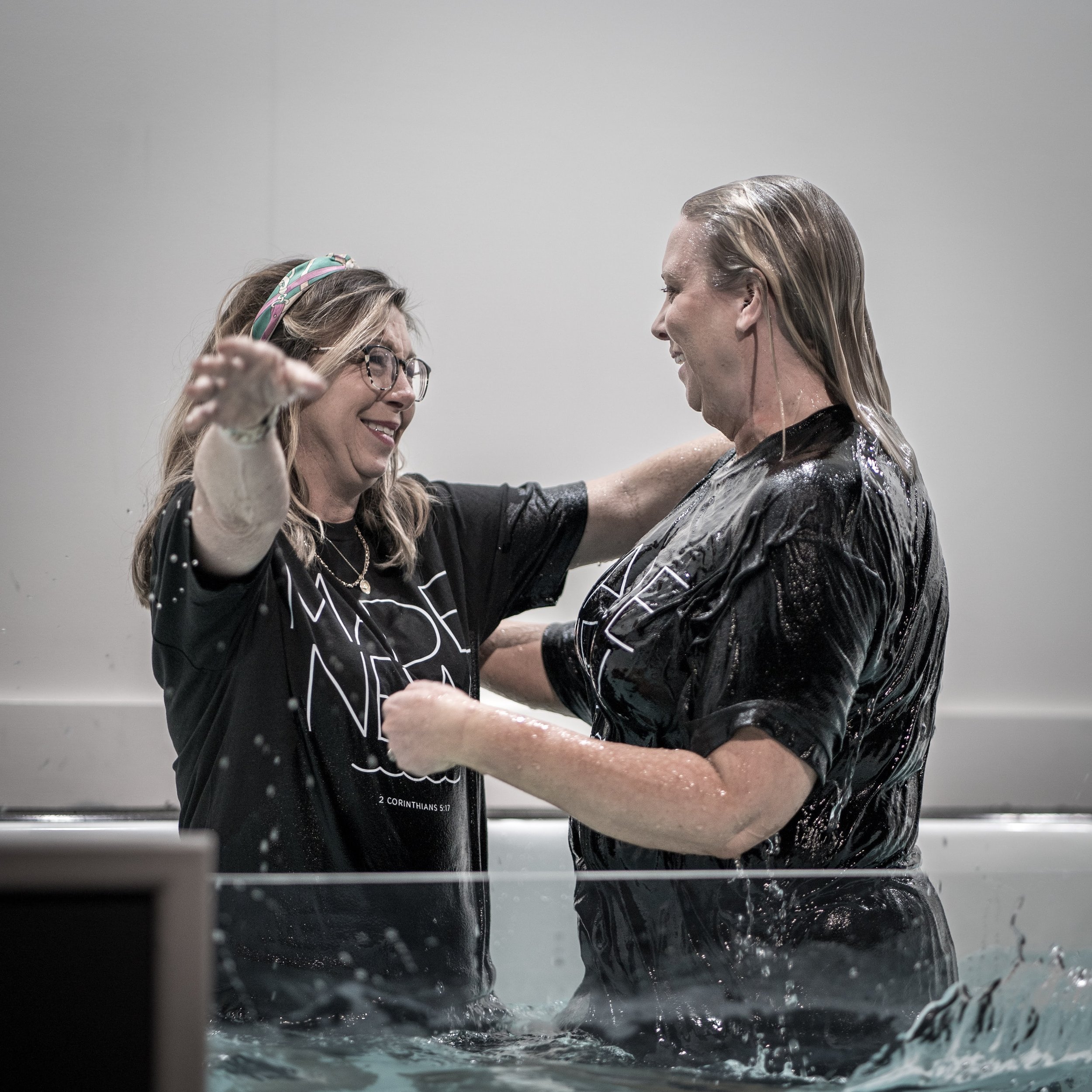 Two women are celebrating in a baptismal pool, one with glasses and a headband, and the other with wet, slicked-back hair, smiling and embracing each other.